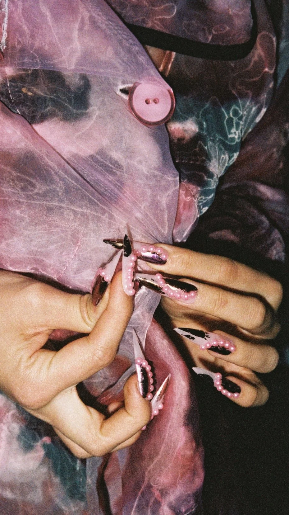 Close-up of hands with long, decorated nails holding a translucent pink fabric with a large button.