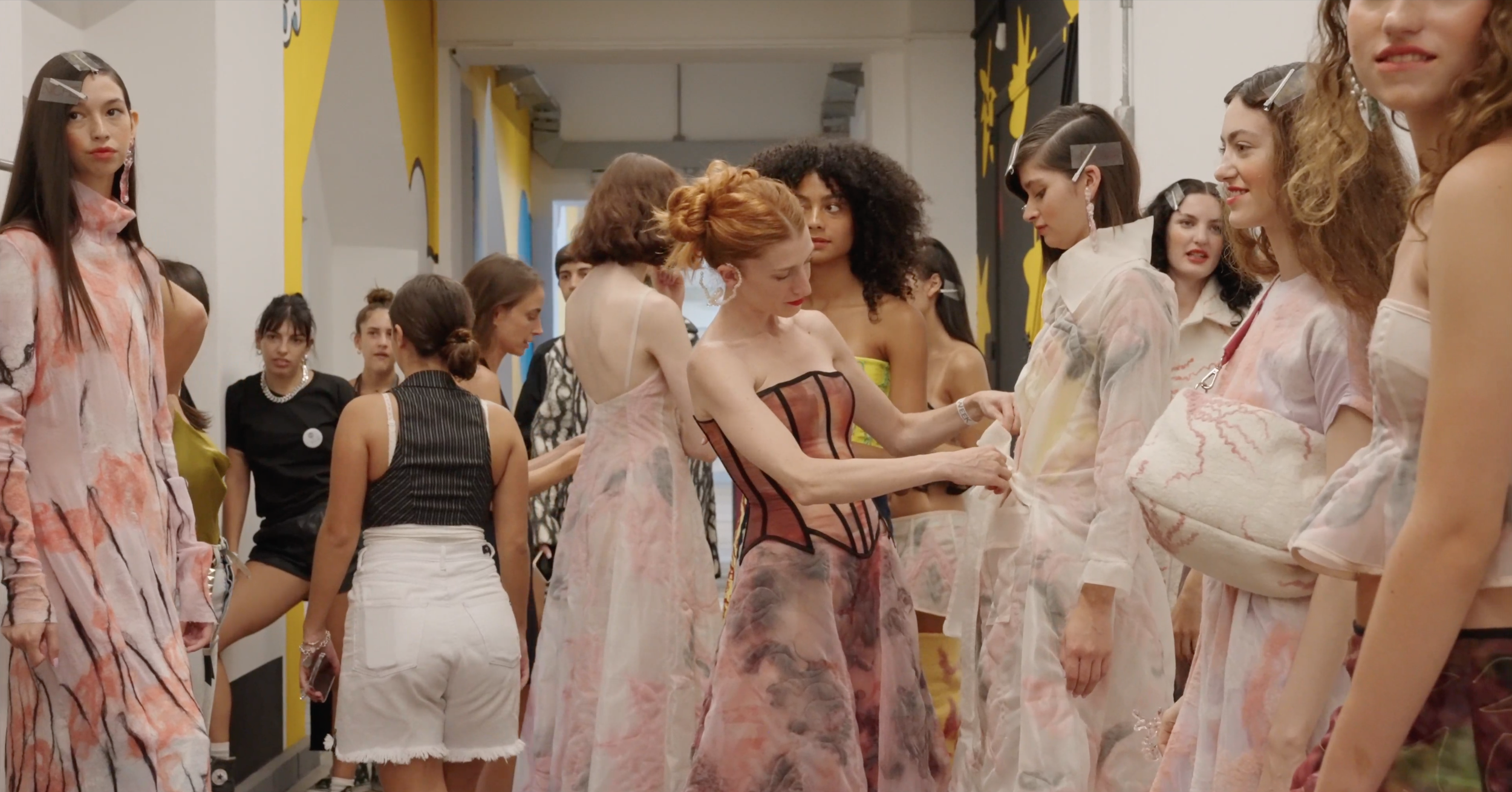Group of young women wearing pastel-colored, abstract patterned dresses in a hallway with yellow and black wall art.