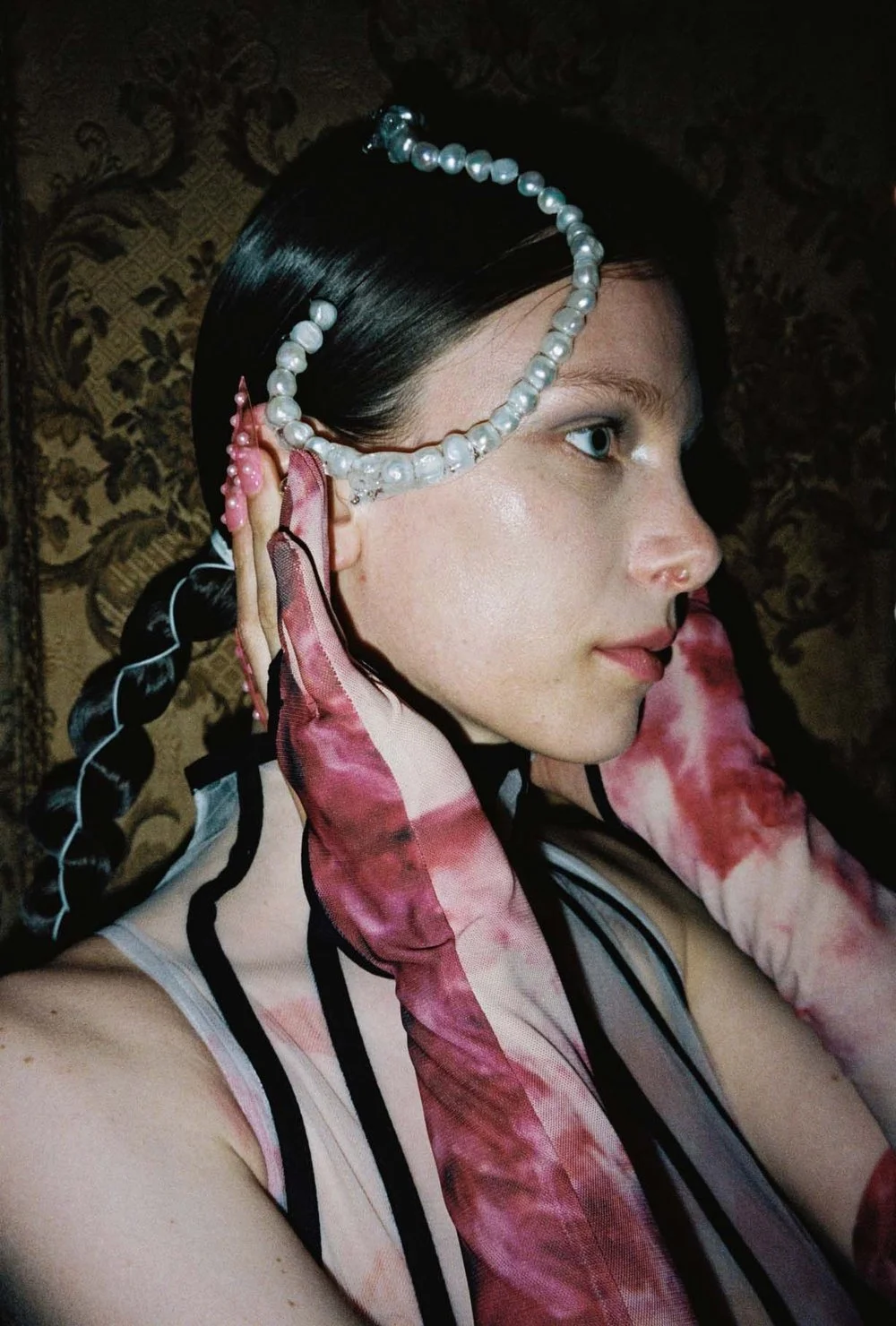 A person with braided hair decorated with pearls, wearing sheer gloves and a patterned top, captured in profile against an ornate background.