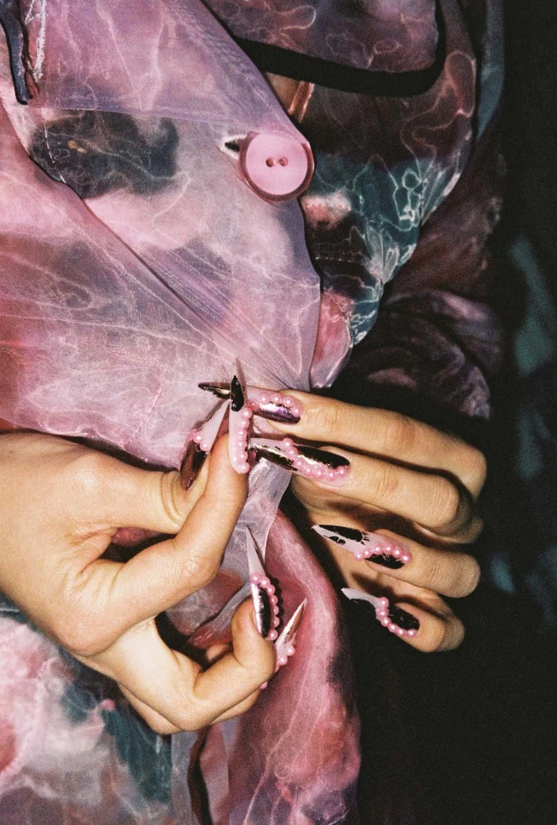 Close-up of hands with long, elaborate pink and black nails, decorated with small pearls, holding a sheer pink and black patterned fabric with a pink button.