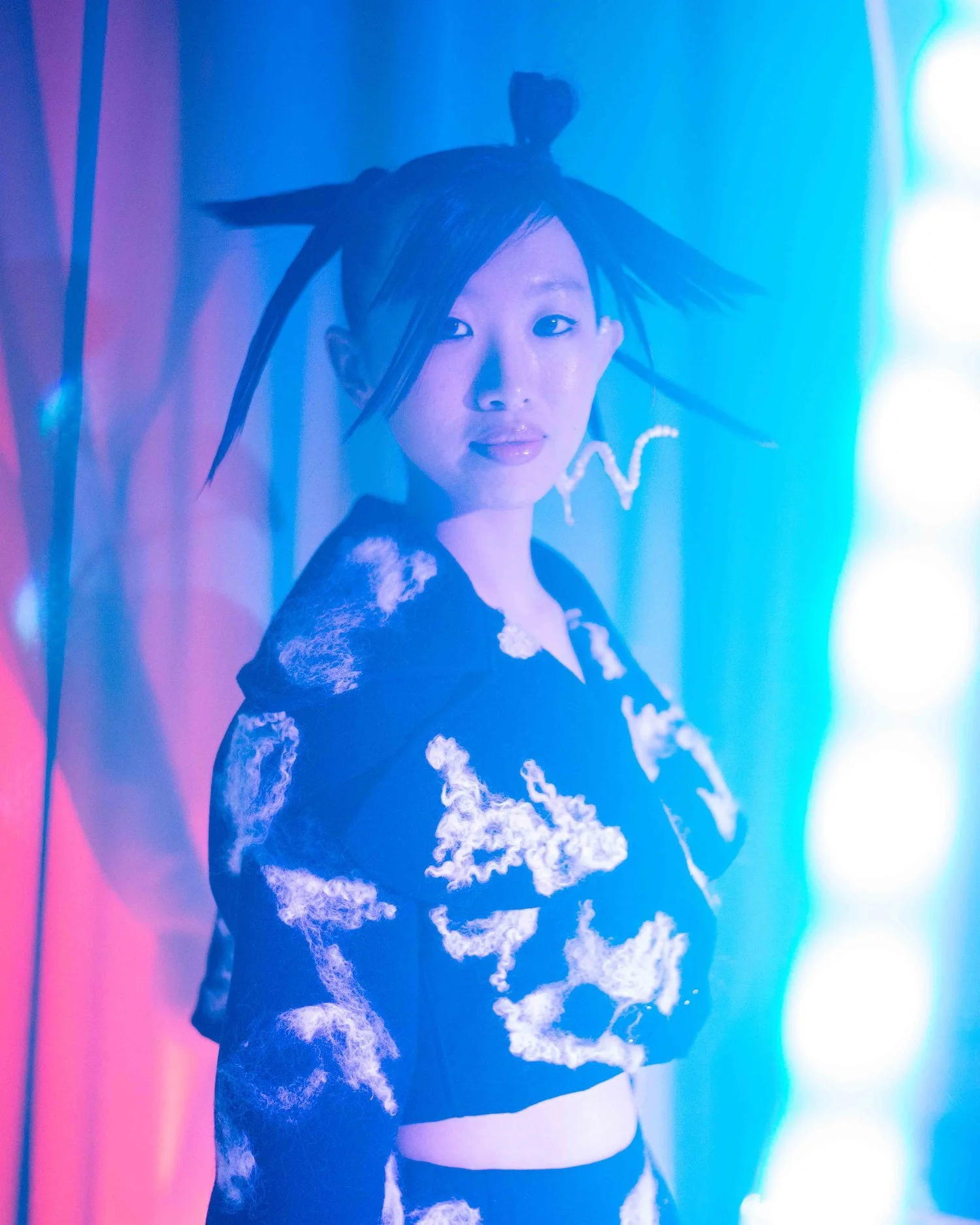 Person wearing a fashionable outfit with abstract patterns in blue and pink lighting, styled hair with unique earrings.