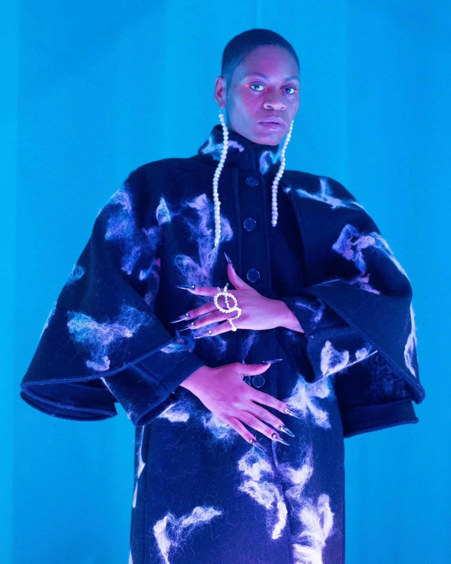 Person wearing a black patterned coat with dramatic pearl earrings and ring, set against a vibrant blue background, expressing a bold fashion statement with intense lighting.