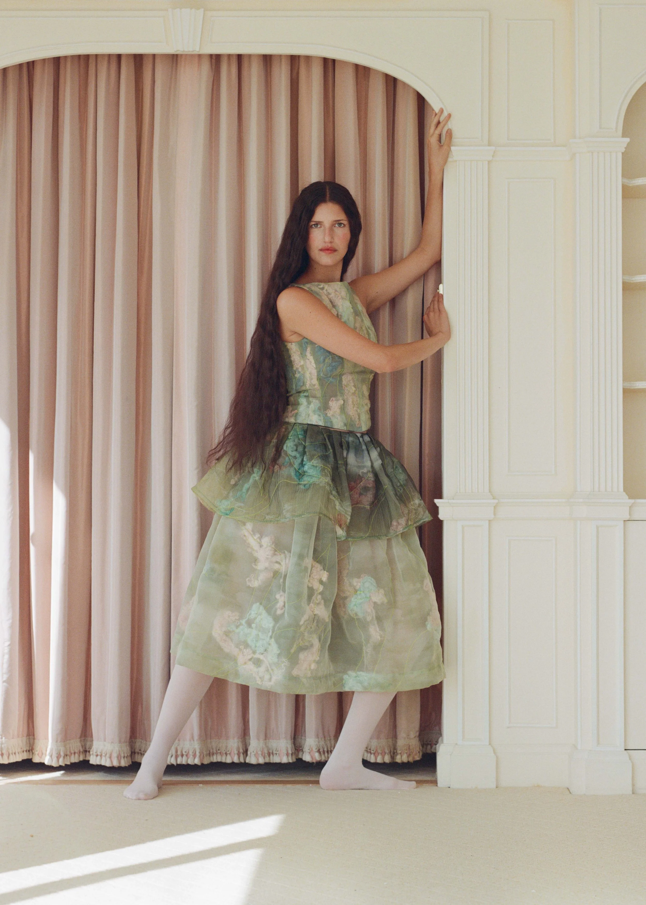A woman with long dark hair standing in front of a pink curtain, holding onto a white column, wearing a green and floral dress with volume, and white tights.
