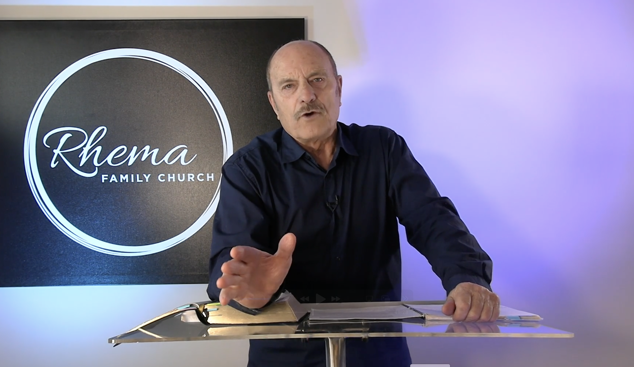 A man with a mustache in a black shirt gestures while speaking, standing in front of a sign that reads 'Rhema Family Church' and a plain wall.