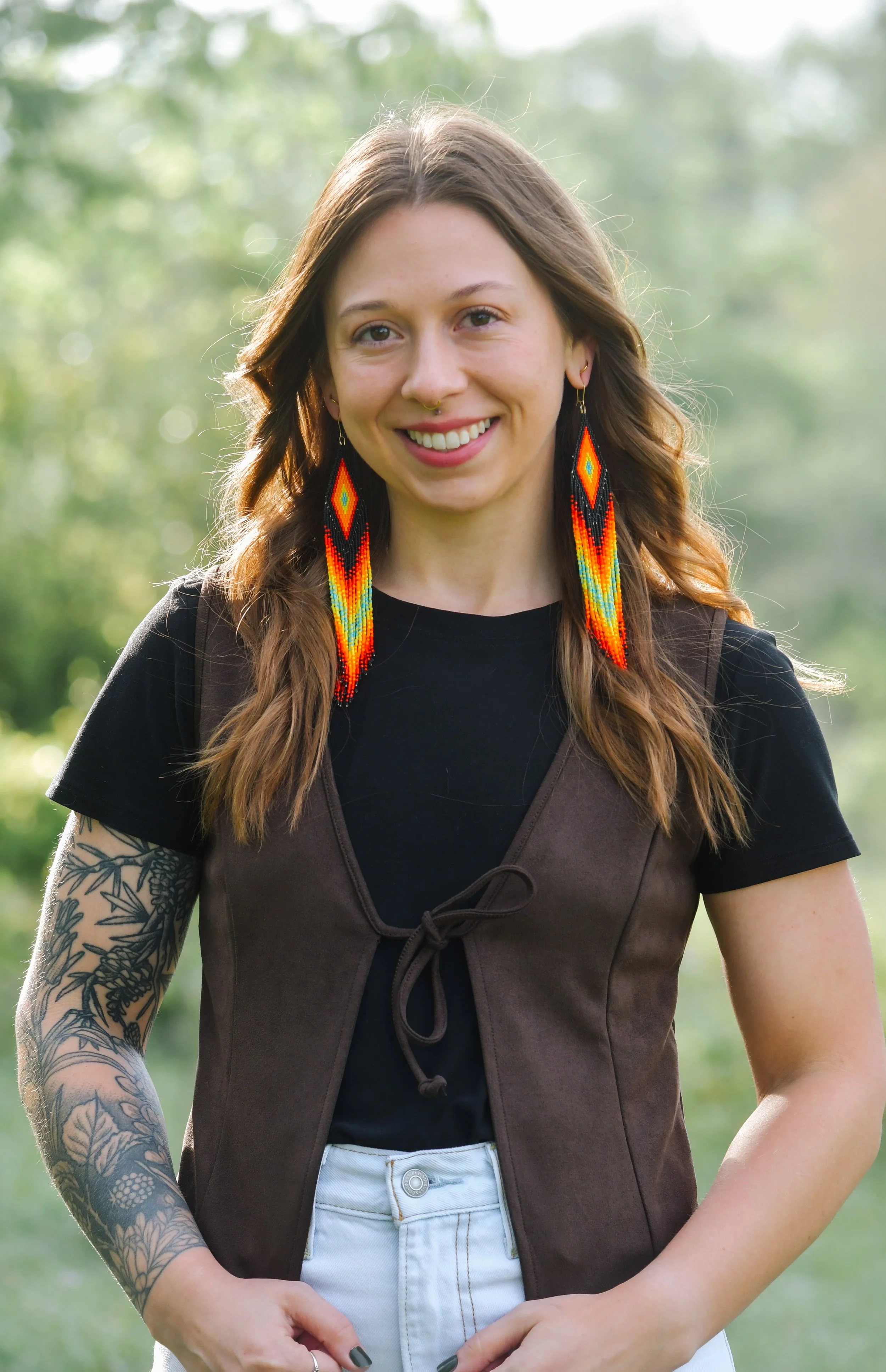 Young woman outdoors with long brown hair, colorful beaded earrings, tattoos on her left arm, wearing a black top with a brown vest, smiling.