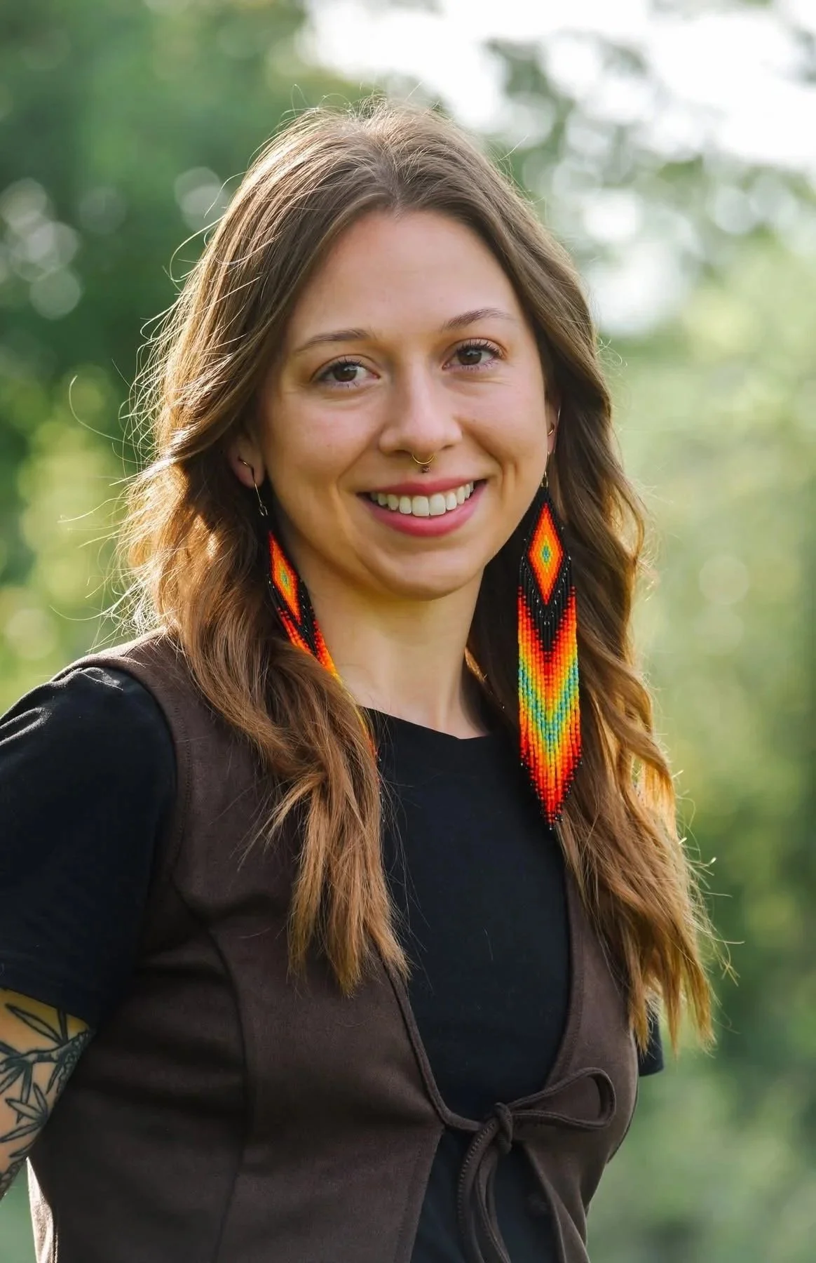 A young woman with long brown hair smiling outdoors, wearing colorful beaded earrings, a black shirt, and a brown vest, with a green blurred background.