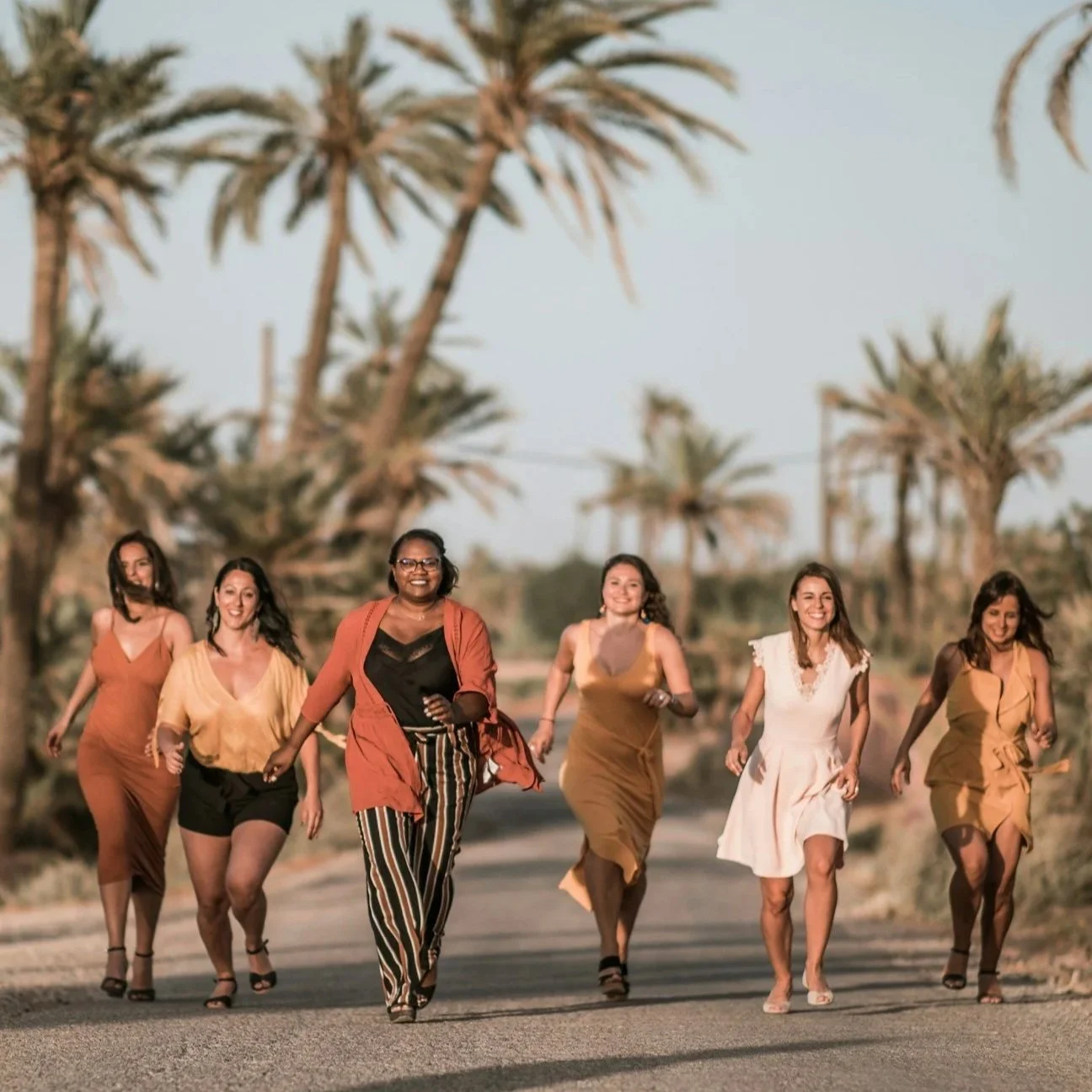 A group of seven women running together on a road, with palm trees and a clear sky in the background. They are smiling and appear to be enjoying themselves.
