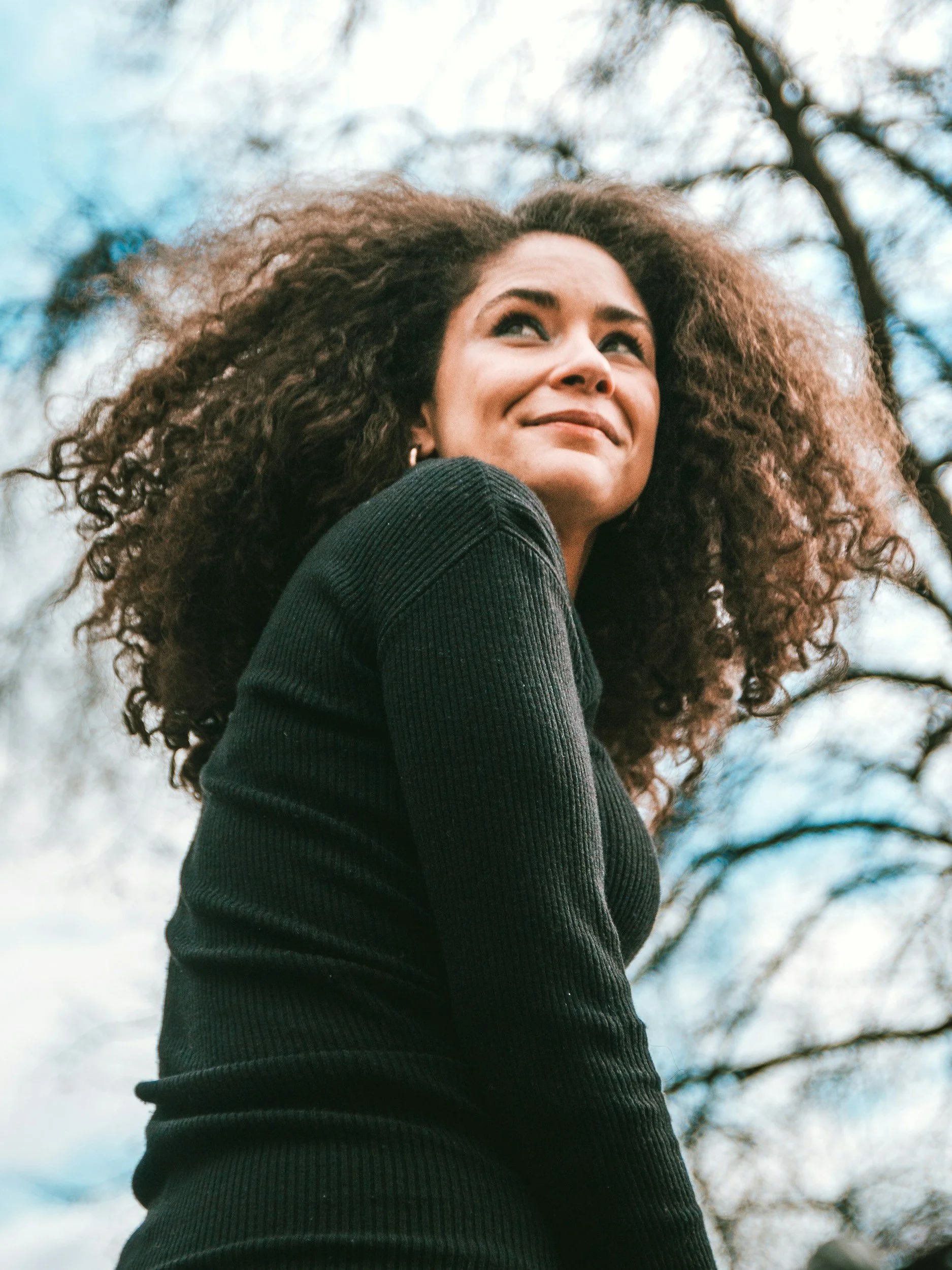 Close-up of a woman with long, curly hair outdoors, smiling slightly, wearing a black sweater, against a background of trees and cloudy sky.