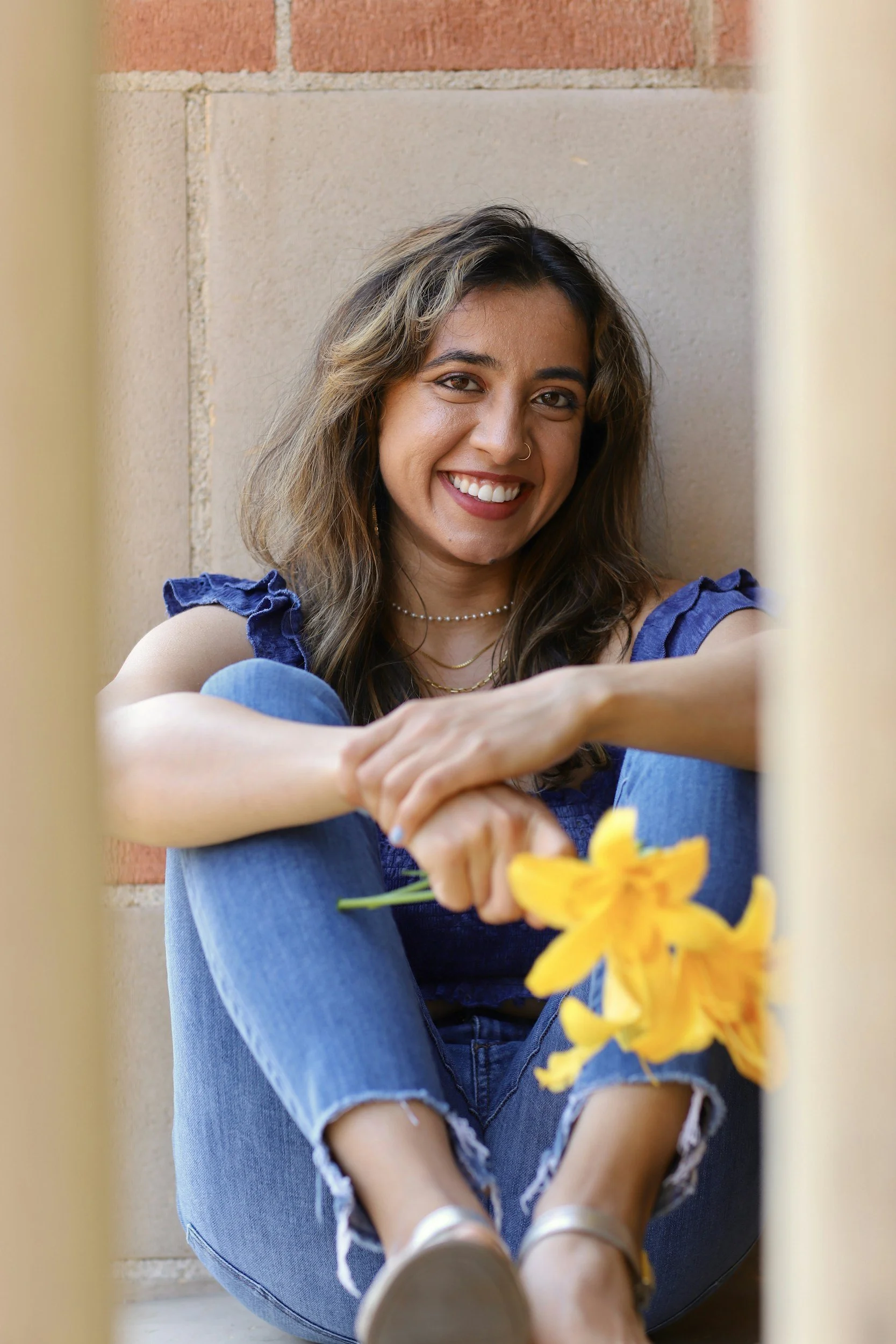 Young woman with wavy hair and a nose ring smiling, sitting against a brick wall, holding a yellow flower.