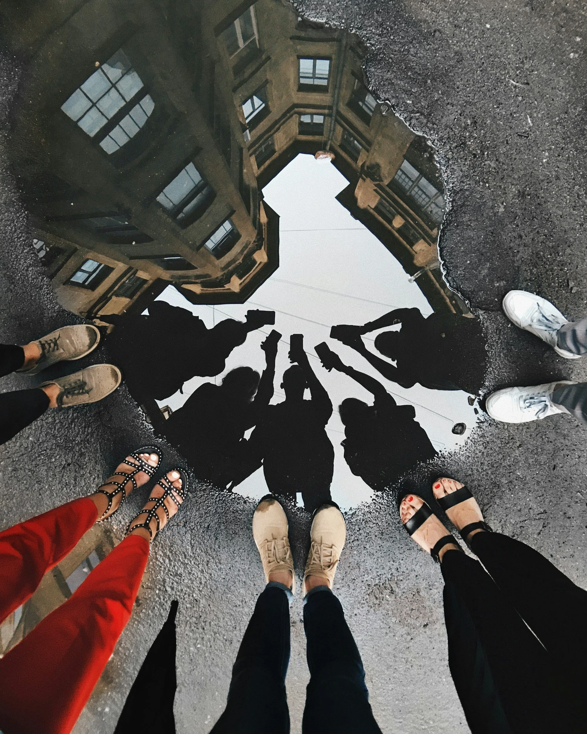 Reflection of a building on a puddle with six people's feet at the edge of the puddle, showing different shoes, with the puddle and building in the background.