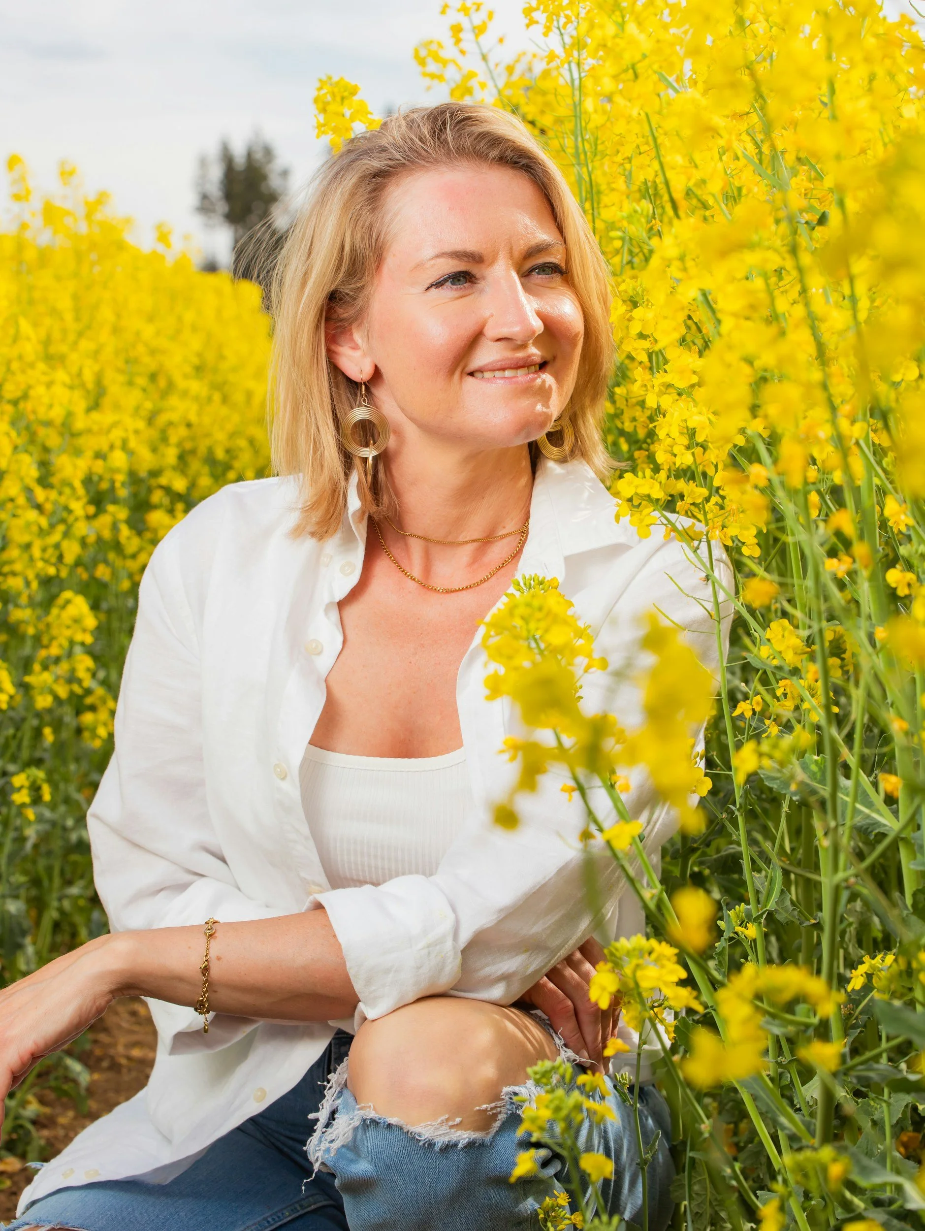 A woman with shoulder-length blonde hair smiling while sitting among yellow flowers in a garden or field.