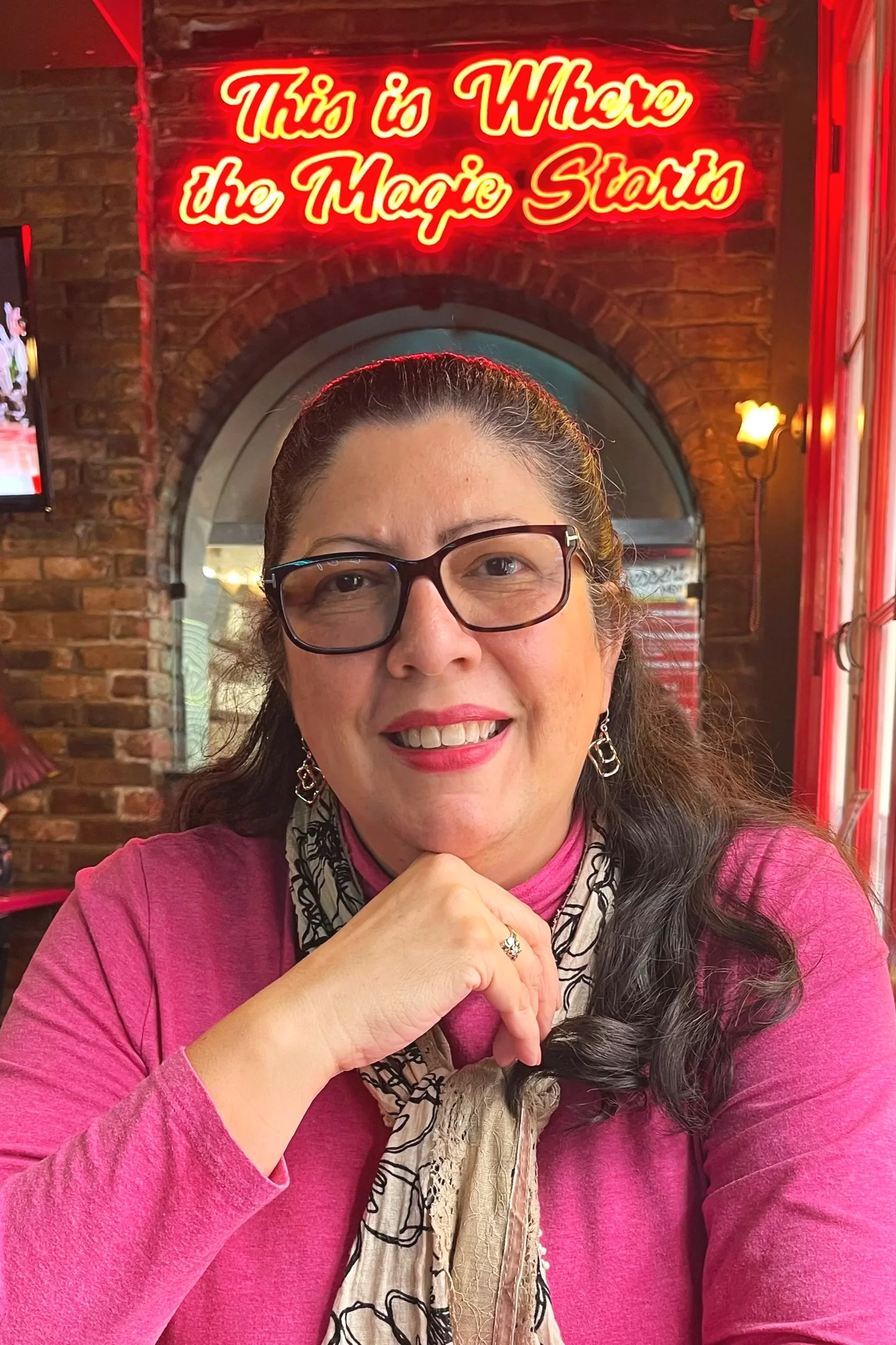 A woman with glasses smiling inside a restaurant with a neon sign that reads "This is Where the Magic Starts". She is wearing a pink top, earrings, and a scarf, and has her hand resting on her chin. The background features exposed brick walls and warm lighting.