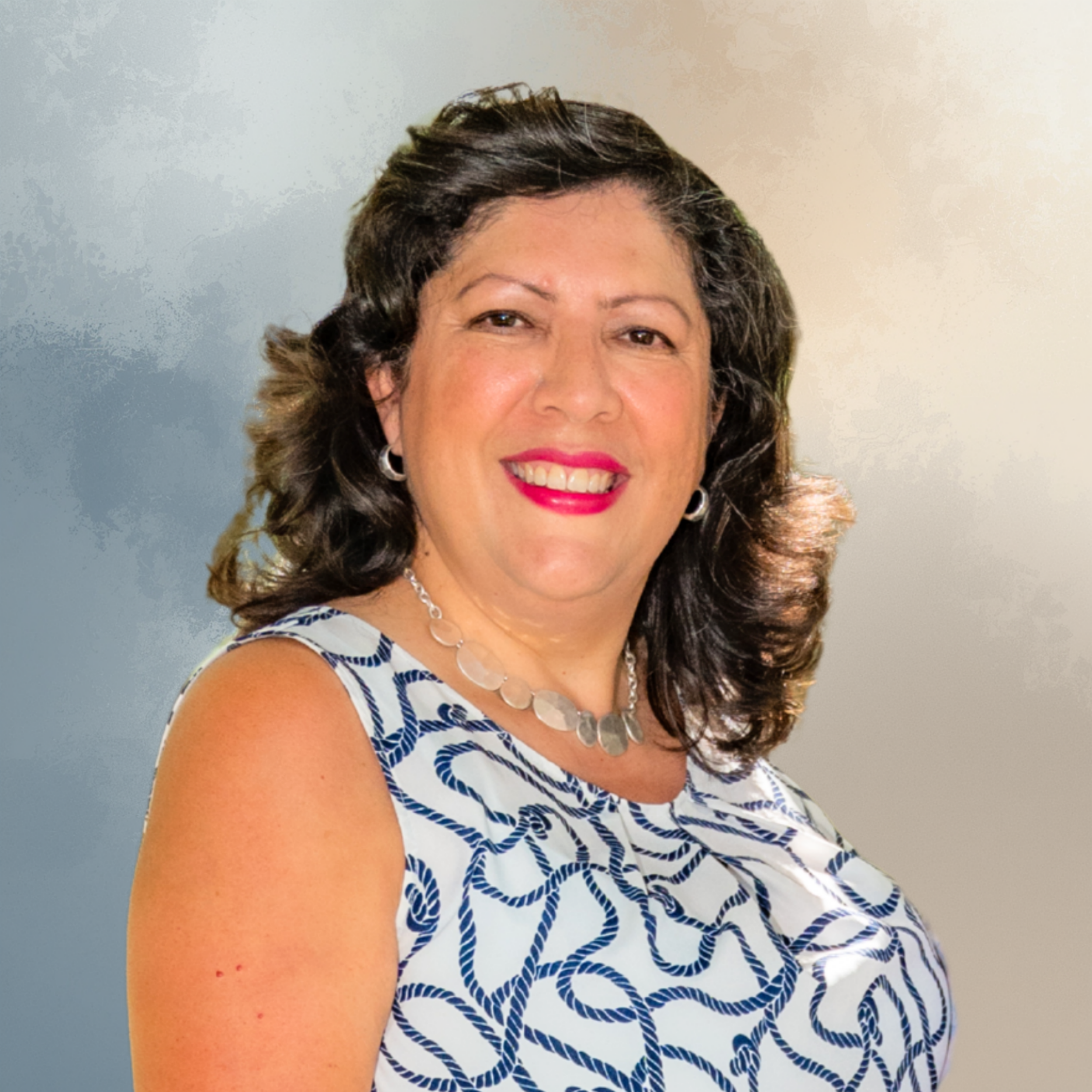 Close-up of a smiling woman with dark, curly hair and earrings, wearing a white dress with blue patterns and a necklace, against a neutral background.