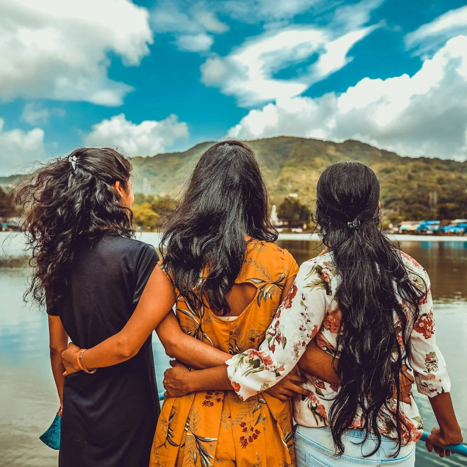 Three women with long dark hair standing arm-in-arm near a body of water, facing a hillside with a partly cloudy sky.
