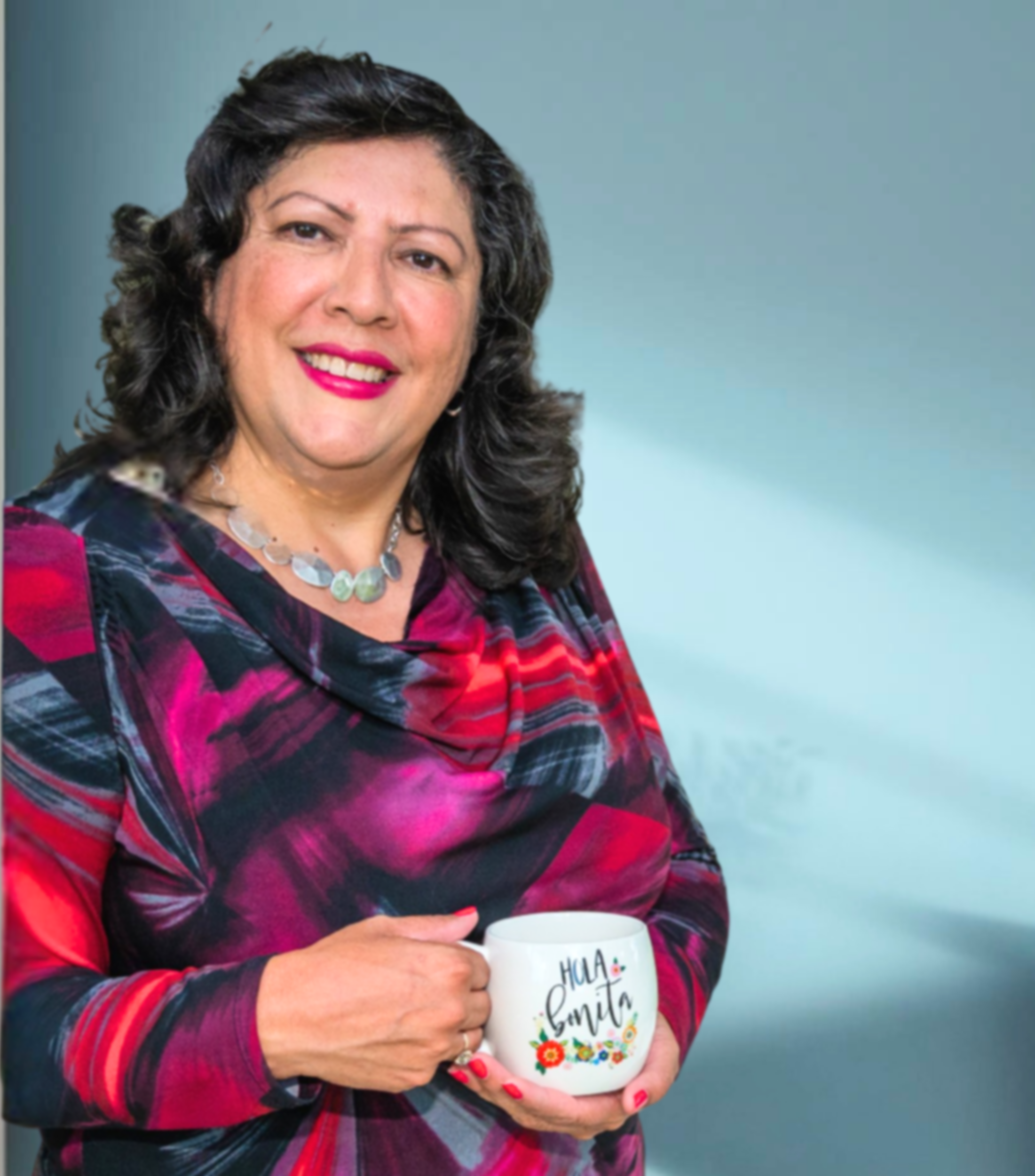 A woman with curly dark hair wearing a plaid shirt and a beaded necklace holding a white mug that says 'Hola Famita' with colorful floral decorations, standing against a plain blue background.