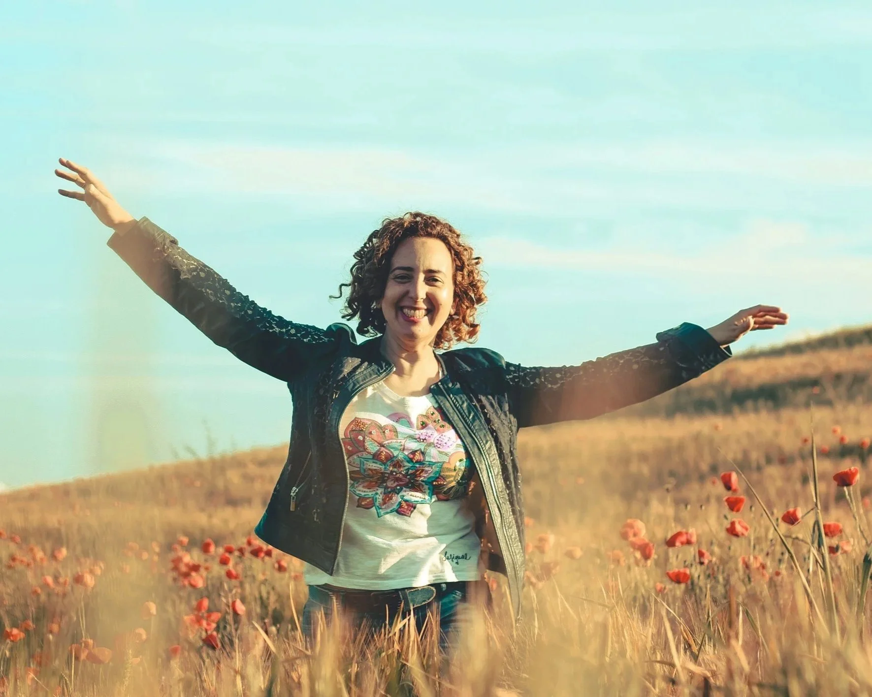 A woman standing in a field of poppies with arms outstretched and a smile, wearing a black leather jacket and a colorful graphic t-shirt, under a bright blue sky.