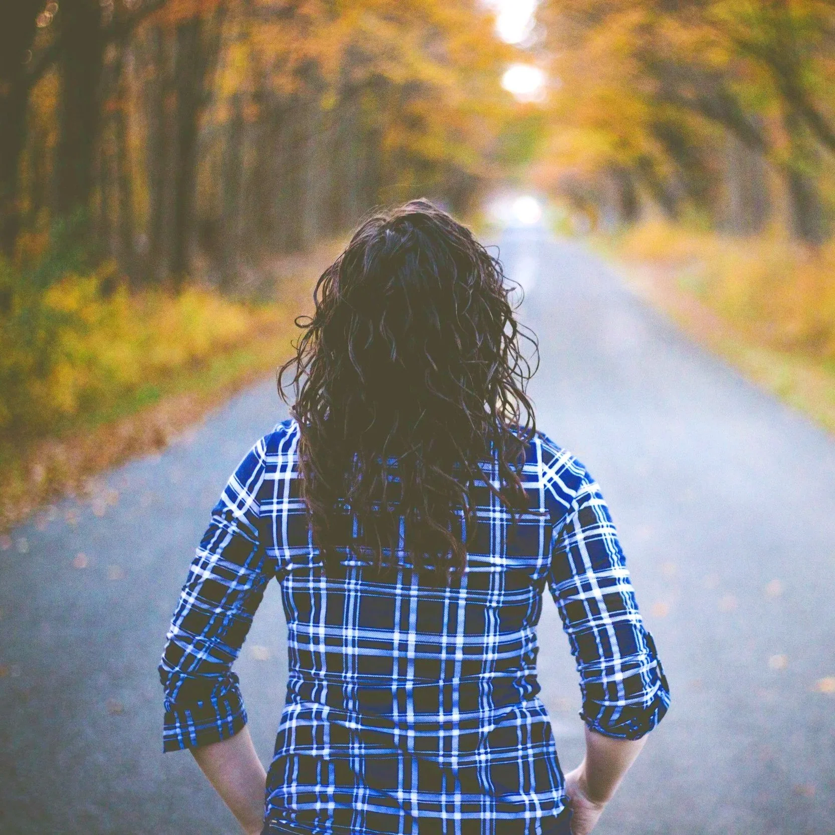 Person with curly hair wearing a blue and white plaid shirt, walking down a tree-lined road in autumn.