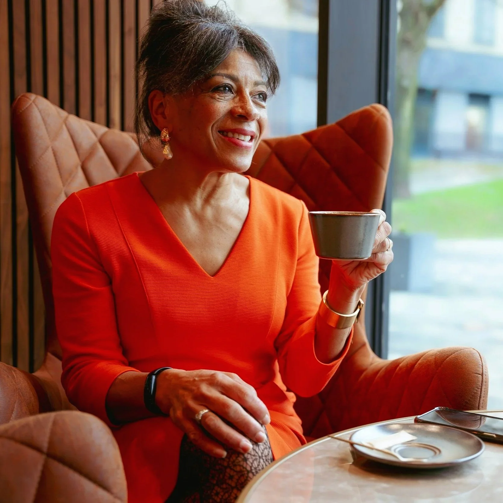 A woman with short gray hair, wearing a red dress, sitting in a cozy cafe by a window, holding a cup, smiling.