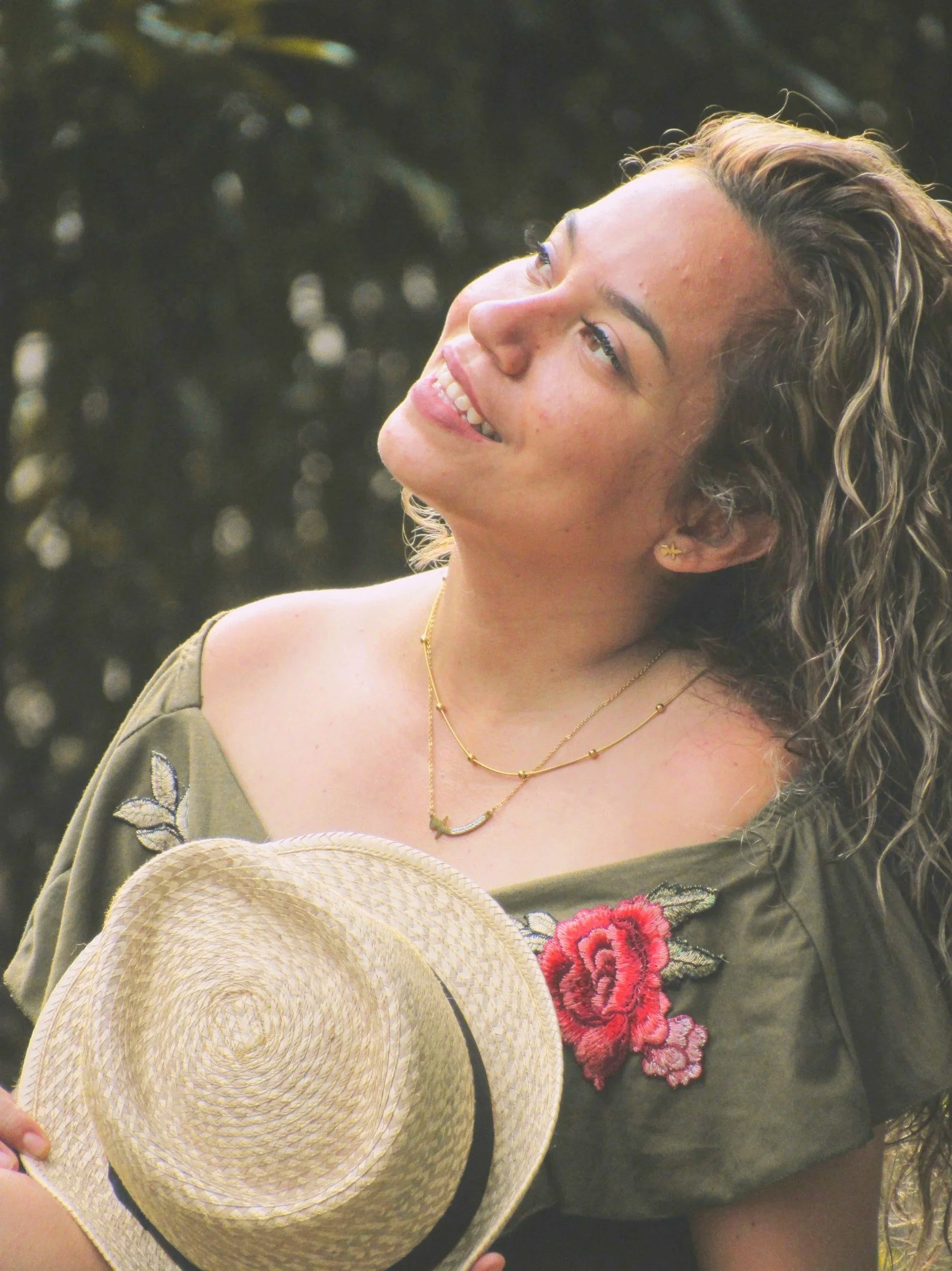 A woman with curly hair smiling and looking up, holding a straw hat, wearing layered gold necklaces and a green dress with floral embroidery.
