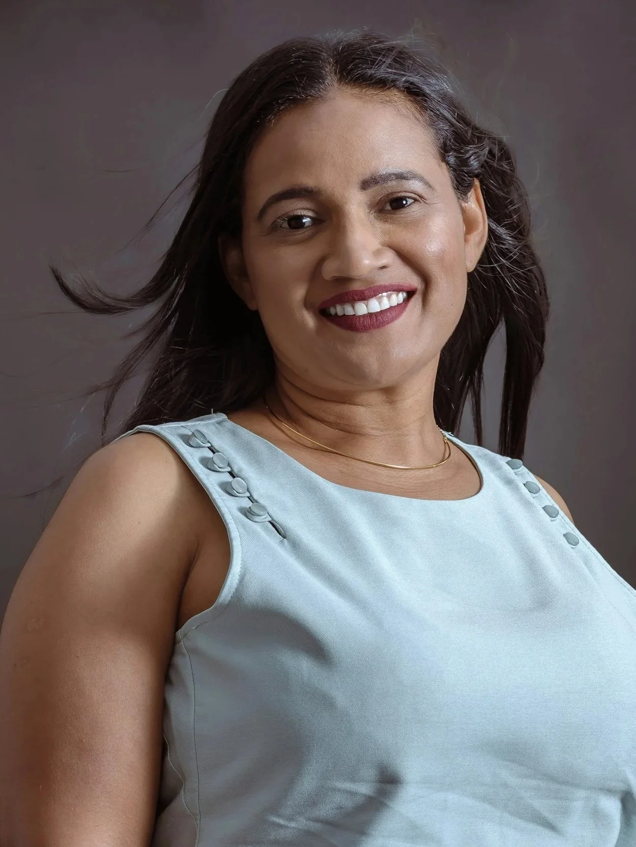 A woman with long dark hair wearing a white sleeveless top with decorative buttons on the shoulders, smiling against a plain gray background.