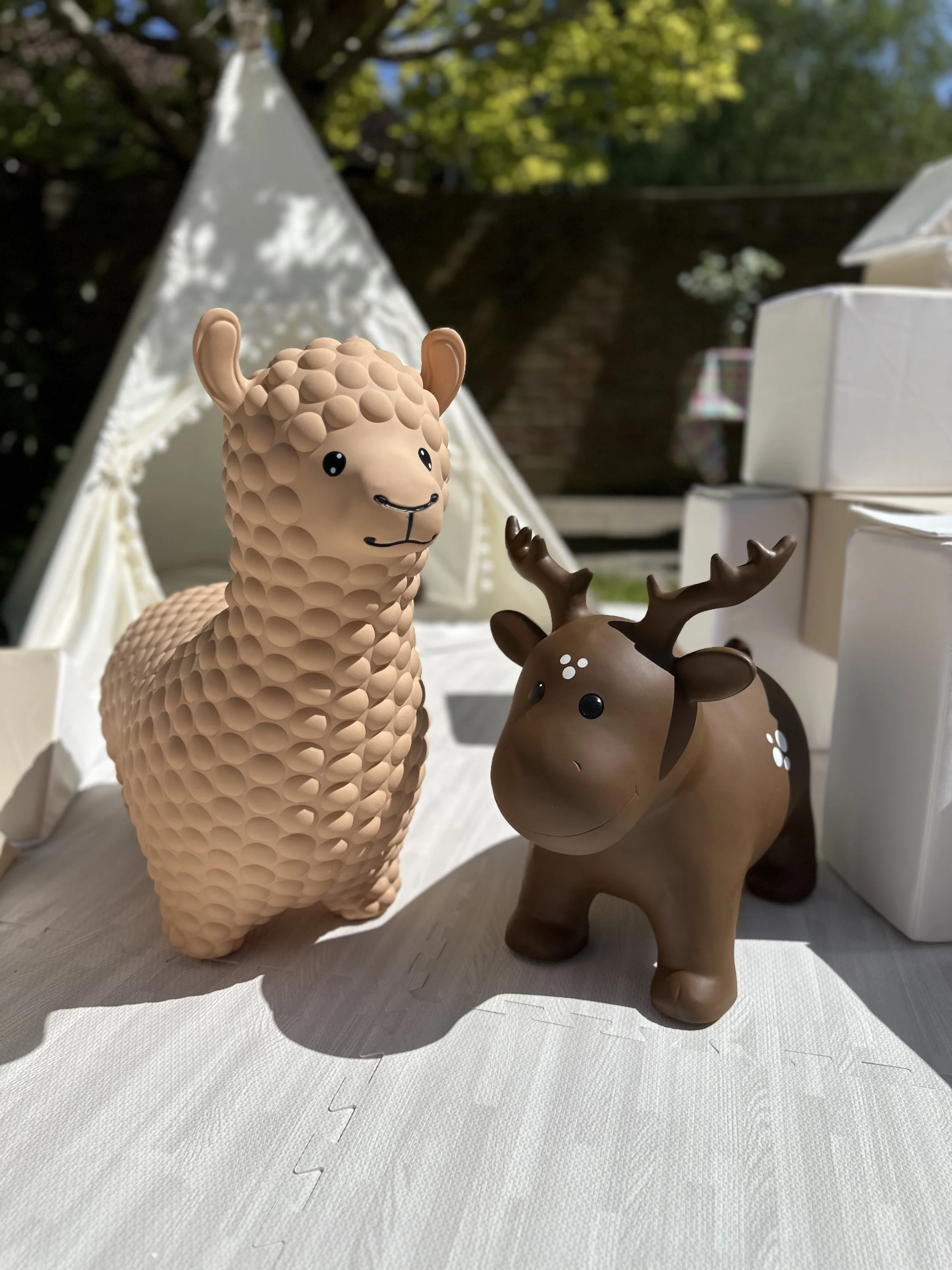 Toy animal figurines, a beige lamb and a brown reindeer, positioned outdoors on a light-colored mat with green trees in the background.