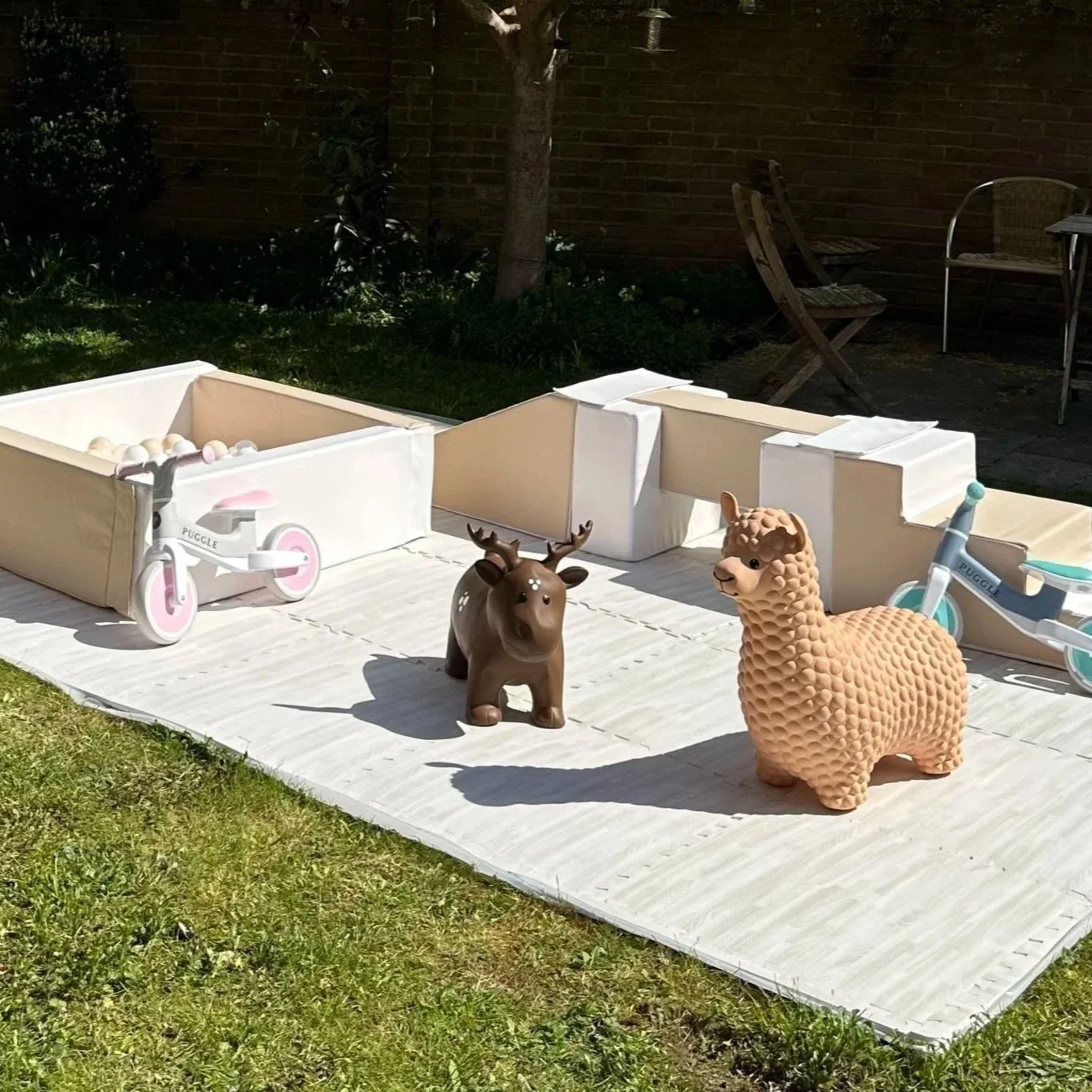 Children's outdoor play area with toy animals and tricycles on a mat, in a backyard with grass, bushes, chairs, and a brick wall.