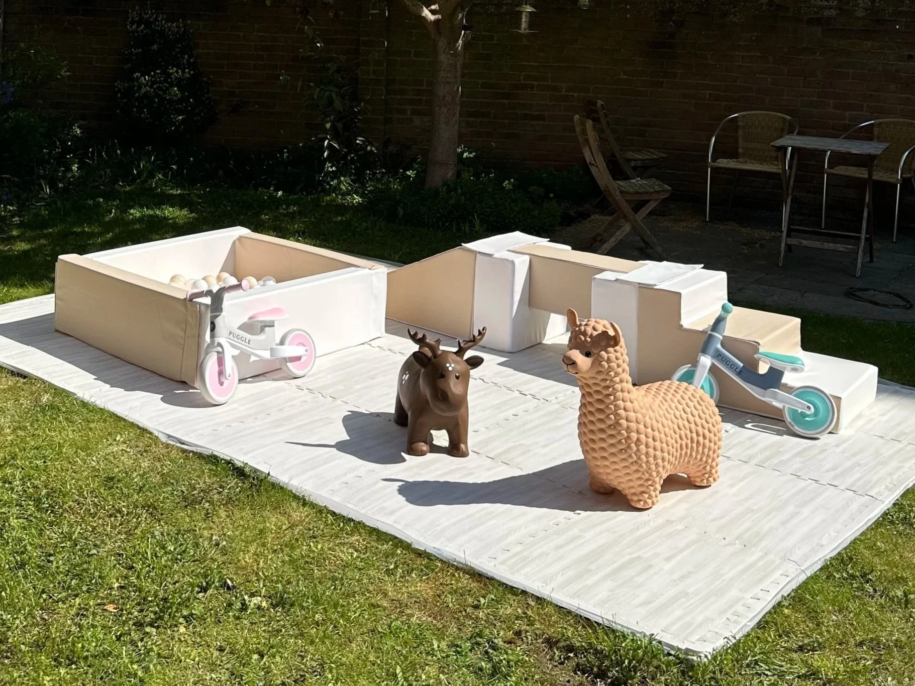 A children's outdoor play area with two toy scooters, one brown moose figure, one beige alpaca figure, on a white mat in a garden with outdoor chairs and a brick wall in the background.
