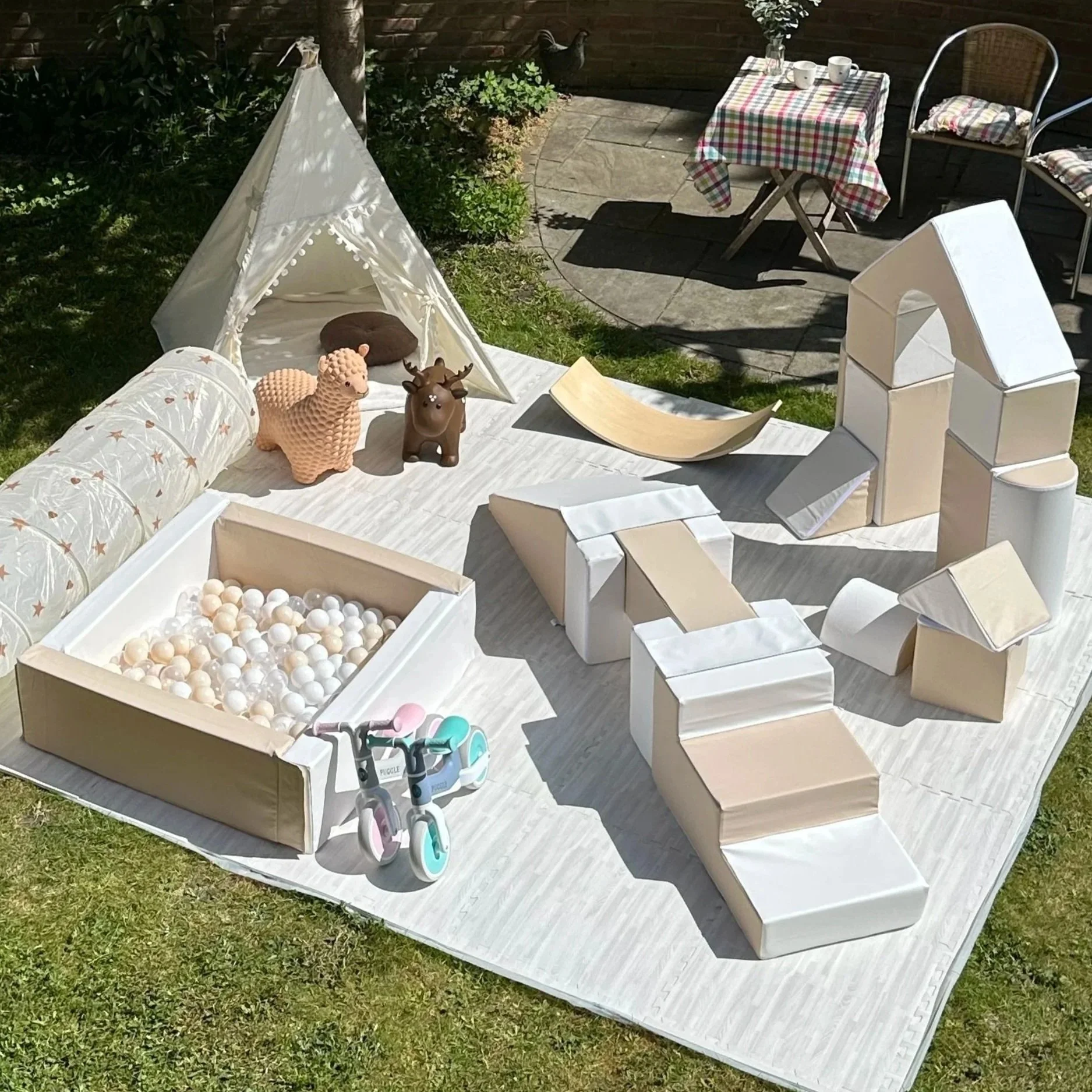 Children's outdoor play area featuring a white teepee tent, a ball pit with white balls, a wooden balance board, a pink toddler bicycle, near a patio surrounded by grass and plants.
