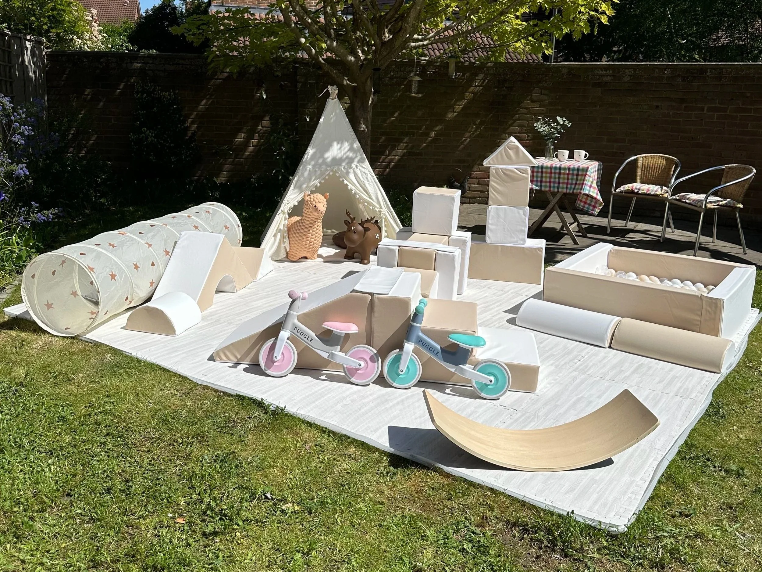 A backyard scene with a small play area featuring a teepee tent, soft blocks, two animal-shaped toys, a toy scooter, and a small table with two chairs. There is also a tablecloth-covered table with cups and a vase of flowers. The area is shaded by a tree, surrounded by plants and a brick wall.
