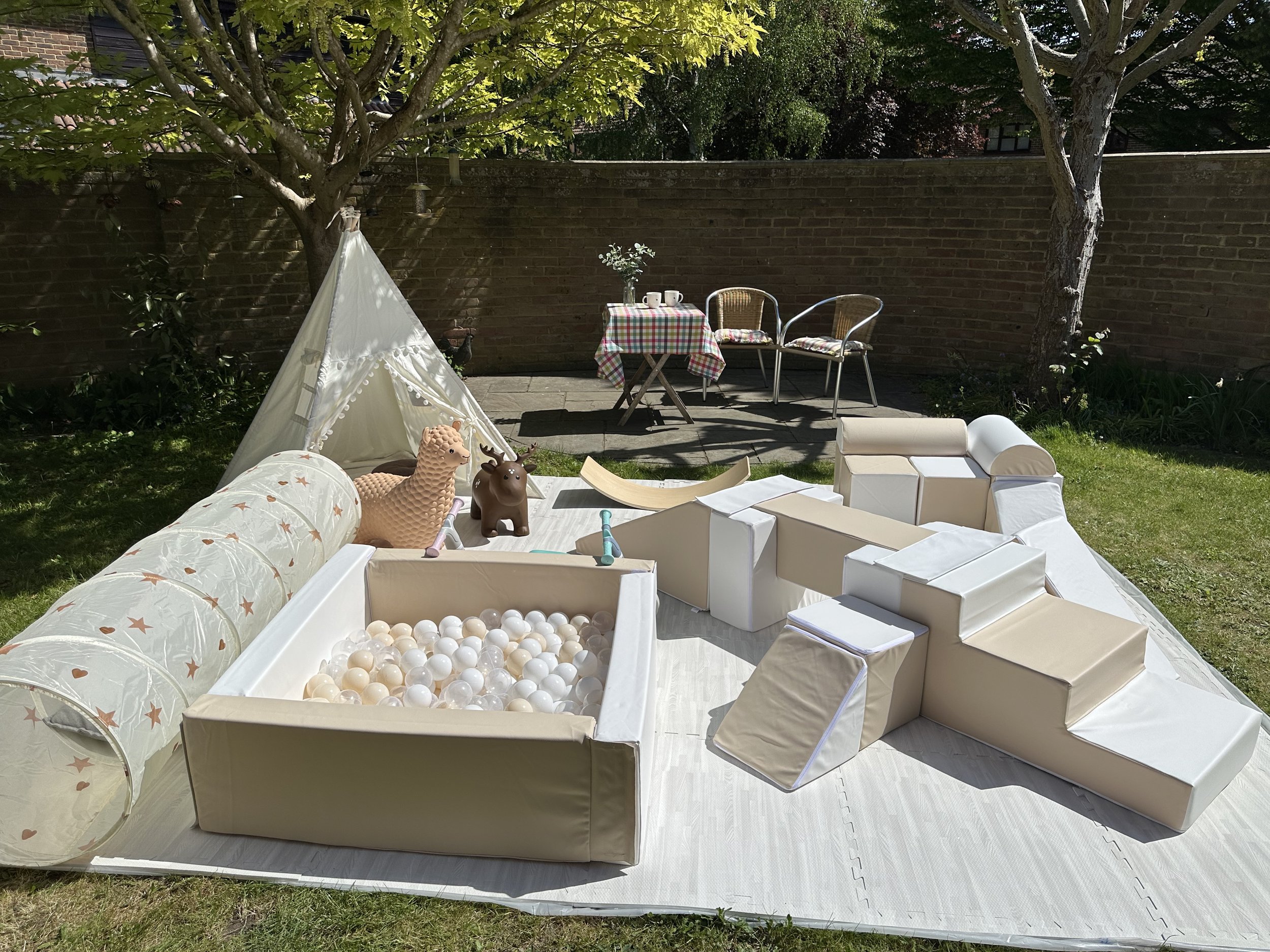 A backyard scene with a small white teepee tent, two animal-shaped toys, a ball pit, and a table with cups and a flower vase, set against a brick wall and surrounded by trees.