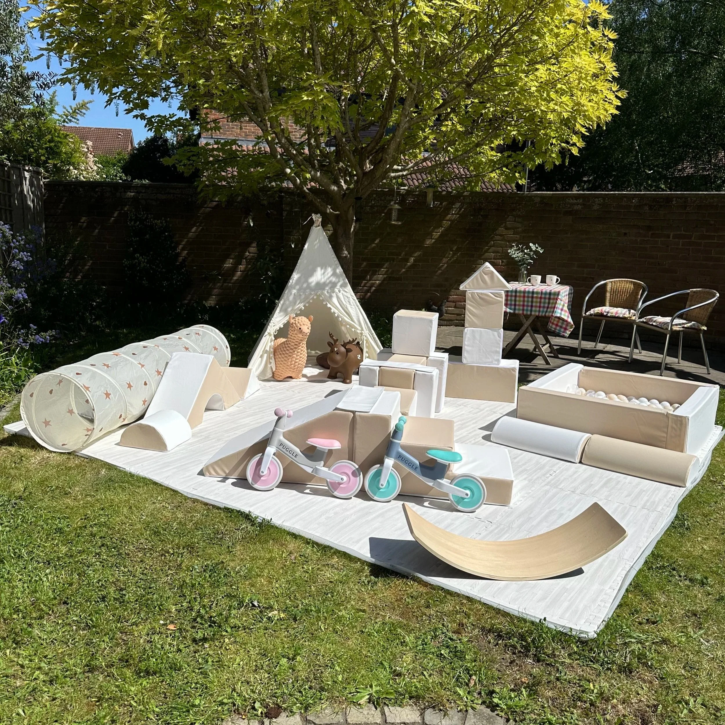 A backyard garden area with a large green leafy tree, brick wall, and some outdoor furniture including chairs, a table with a checkered tablecloth, and children's toys like a tricycle and animal figurines. The scene is illuminated by sunlight.
