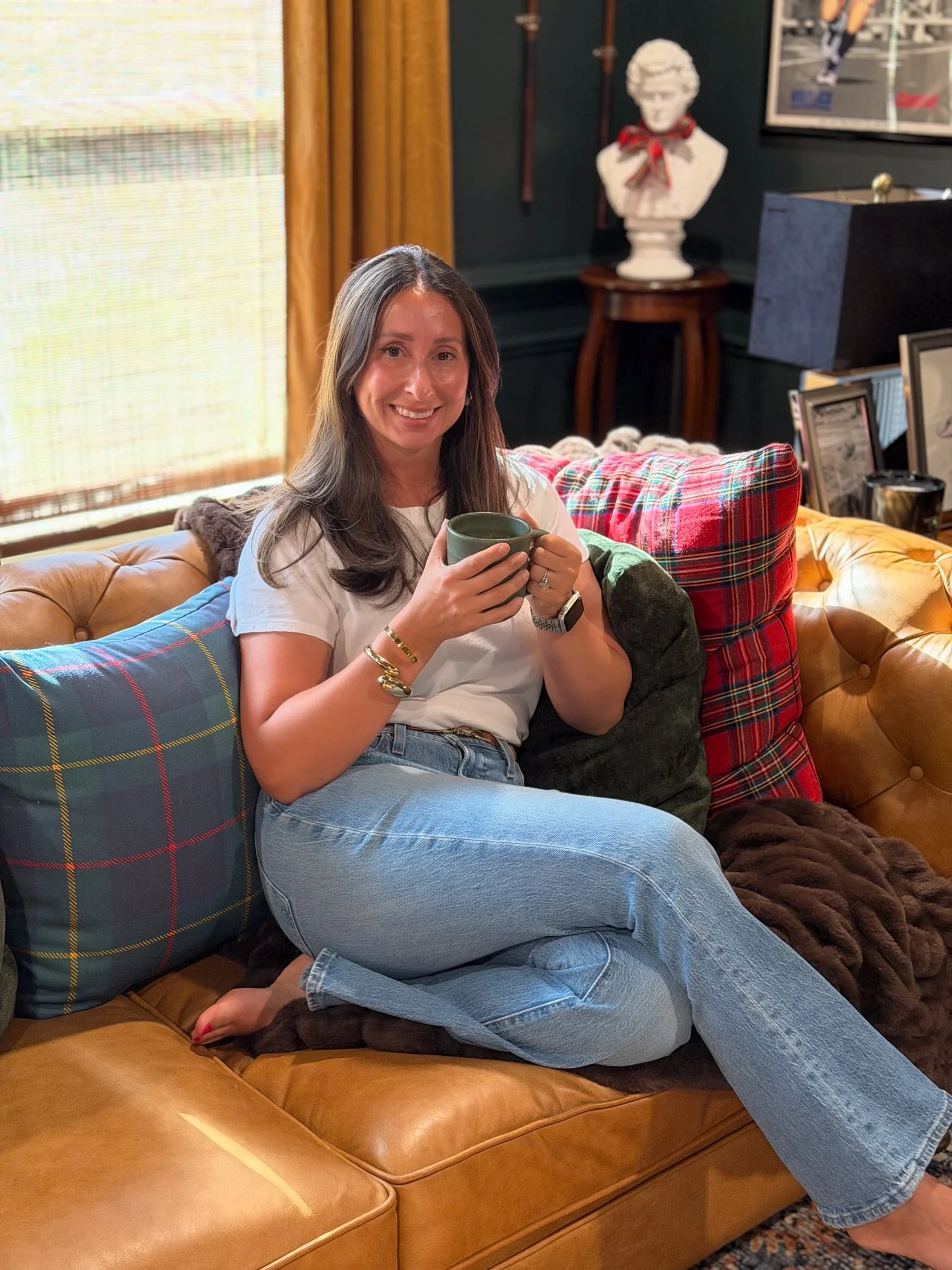 Woman sitting on a tan leather sofa, holding a green mug, smiling. She has long dark hair, wearing a white t-shirt and blue jeans. The sofa is decorated with plaid and solid colored cushions. In the background, there are framed pictures, a marble bust with a red bow, and a window with beige curtains.