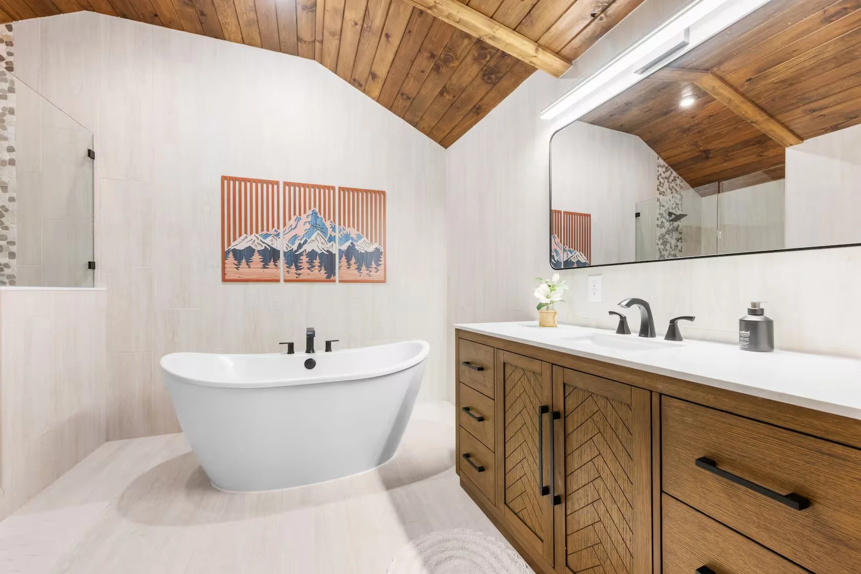 Modern bathroom with a wooden vaulted ceiling, a white freestanding bathtub, a large mirror above a wooden vanity with black hardware, a small potted plant, and mountain-themed artwork on the wall.