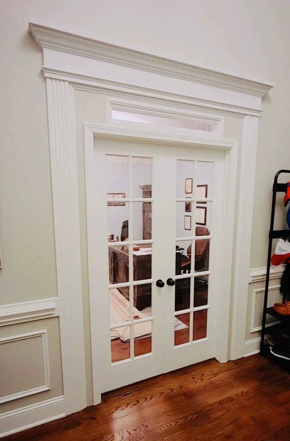 Interior view of a set of white double French doors with glass panes, decorative crown molding, and chair rail molding in a room with hardwood floors.