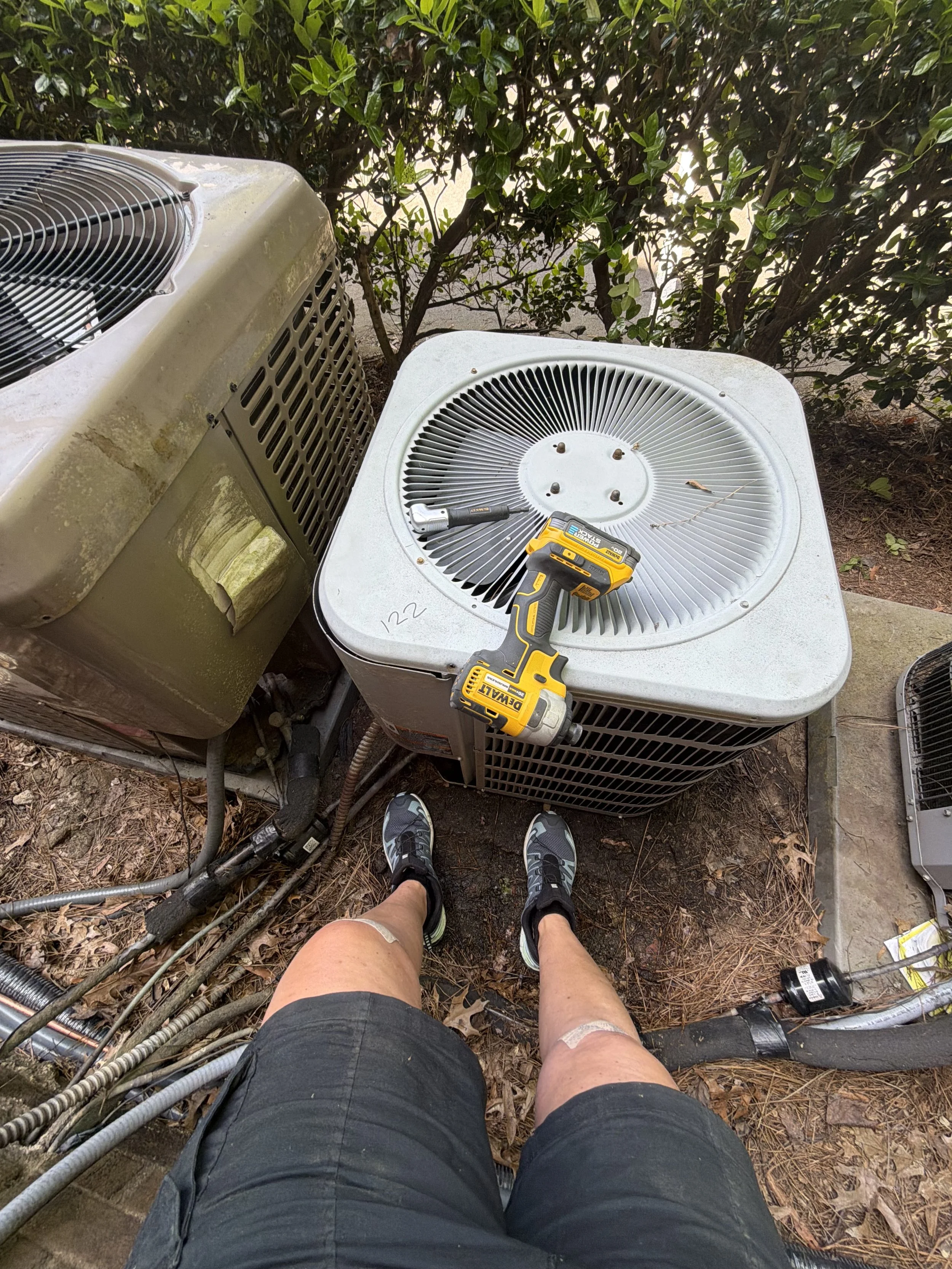 A person wearing black shorts and sneakers standing outdoors on a dirt ground with leaves, surrounded by old outdoor air conditioning units, with tools and wires resting on top of one unit, and a tree with green leaves in the background.