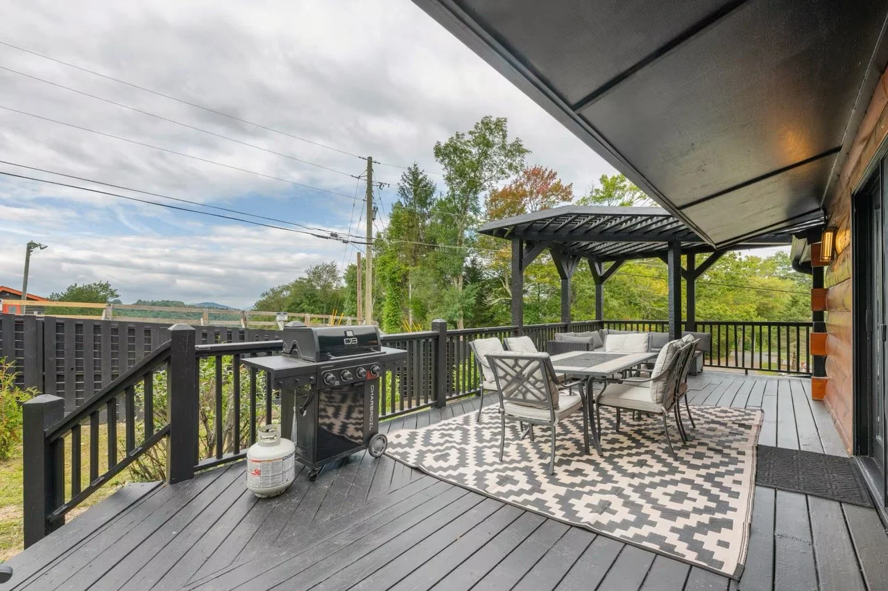Outdoor deck with black railing, covered area with patio furniture, a dining table with eight chairs, a black gas grill, outdoor rug, propane tank, and a scenic view of trees and cloudy sky in the background.