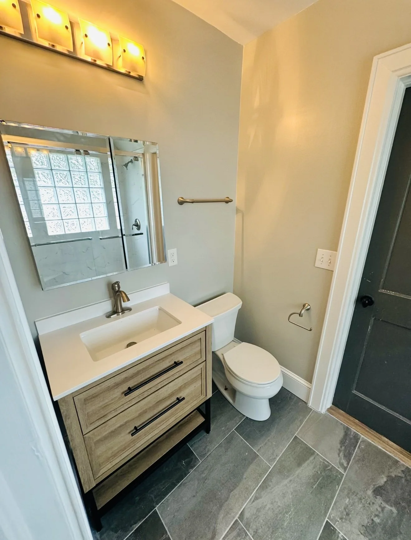 A small bathroom with a modern design, featuring a wooden vanity with black handles, a white sink, a mirror, a toilet, a towel bar, and a glass shower enclosure, with gray tile flooring and light-colored walls.