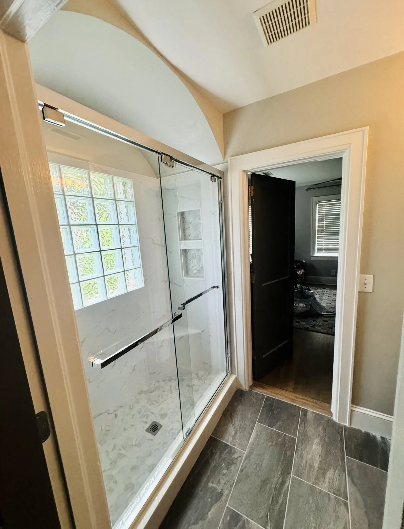 Bathroom with a glass shower enclosure, marble tiles, window made of glass blocks, and an open doorway leading to another room with a window and patterned rug.