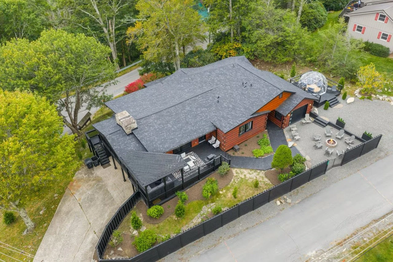 Aerial view of a house with a dark gray roof, red-brown wooden walls, outdoor deck, firepit area with chairs, a geodesic dome, and surrounding trees and neighborhood.