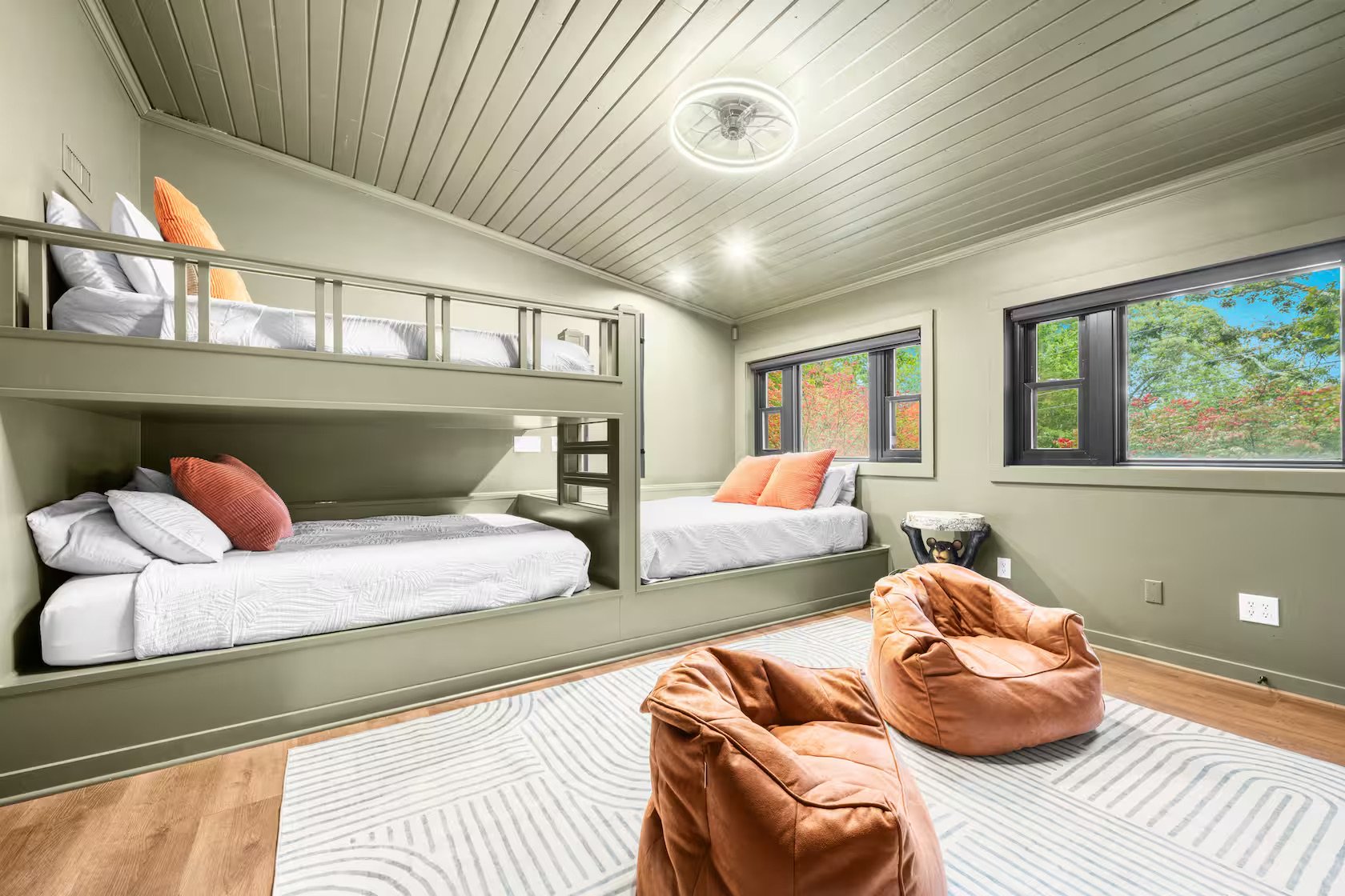 A bedroom with two beds, one on the floor and one on a bunk bed, with white and orange pillows. The room has three windows showing greenery outside, a ceiling fan or light fixture, and a corner side table with a decorative item. Two bean bag chairs are placed on a striped rug on the wooden floor.