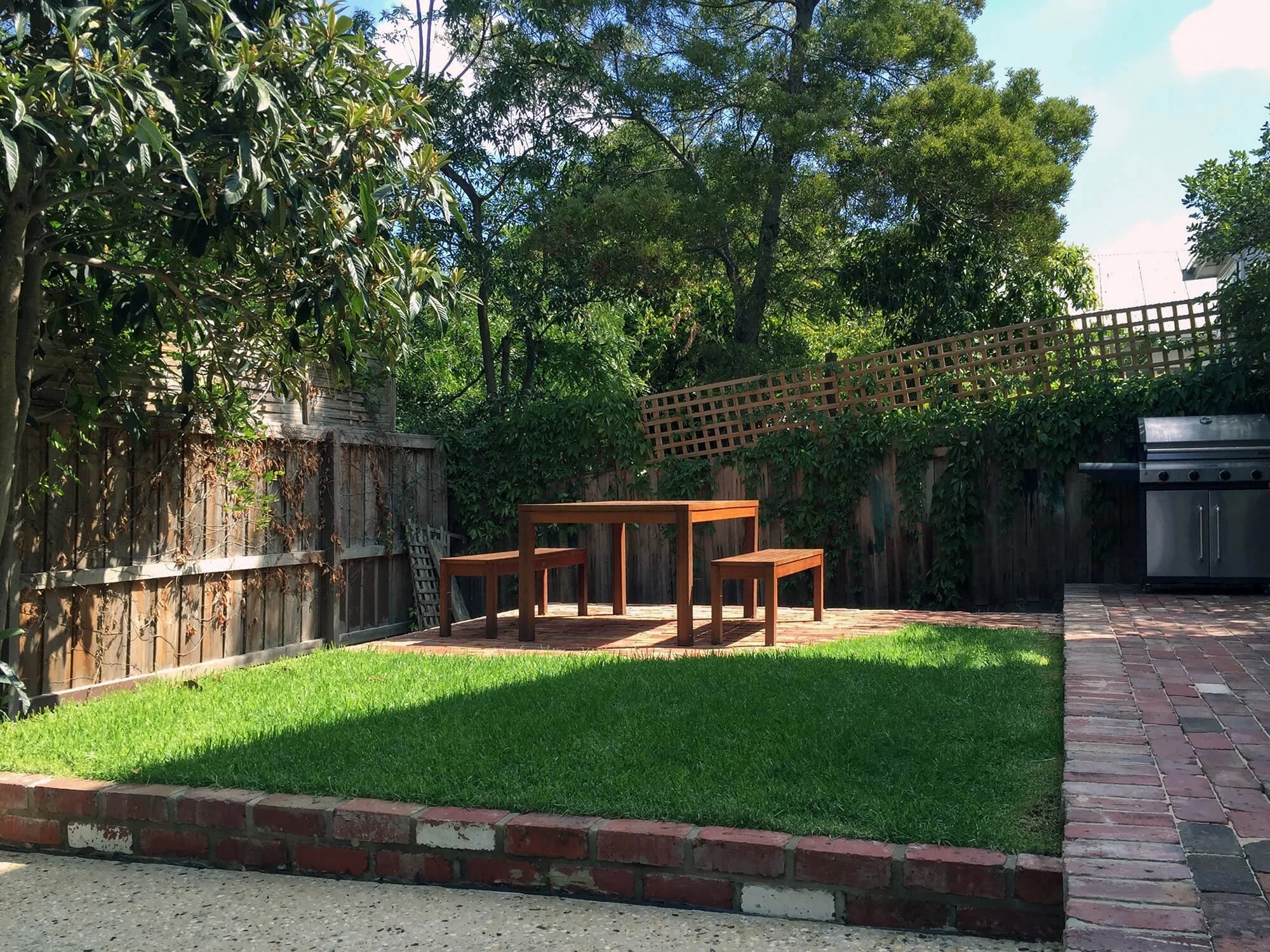 A backyard with a brick patio, a lush green lawn, a wooden table and chairs, and a tall fence surrounded by dense trees and shrubs under a partly cloudy sky.