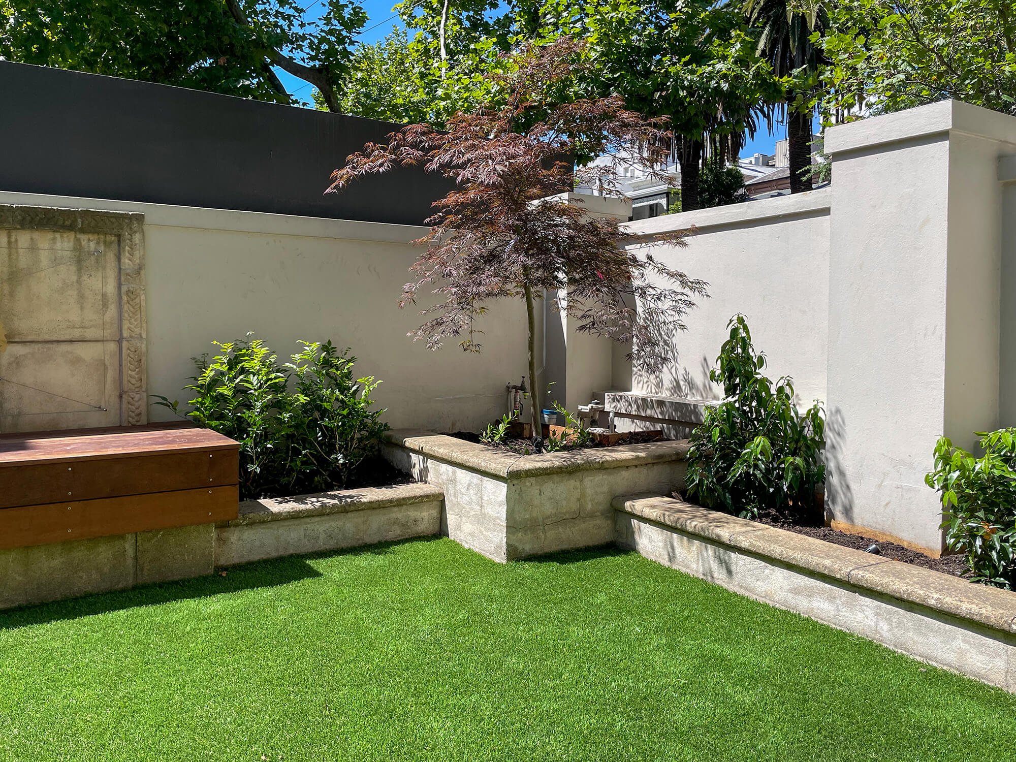 A backyard with artificial green grass, a concrete and wood raised garden bed with various plants, a small tree with reddish leaves, a white stucco wall, and green trees and blue sky in the background.