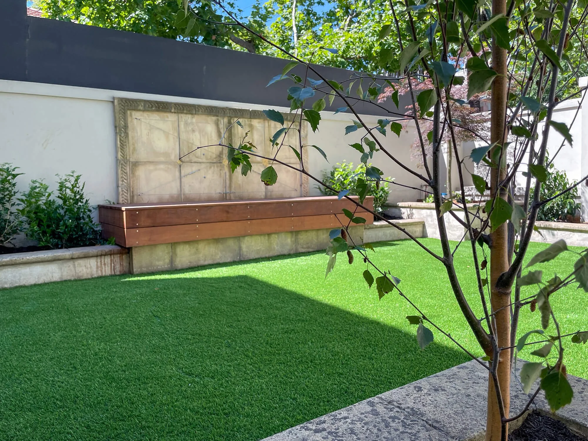 A backyard garden featuring a tree with green leaves, a wooden planter box, green grass, and a concrete pathway with a wall and trees in the background.