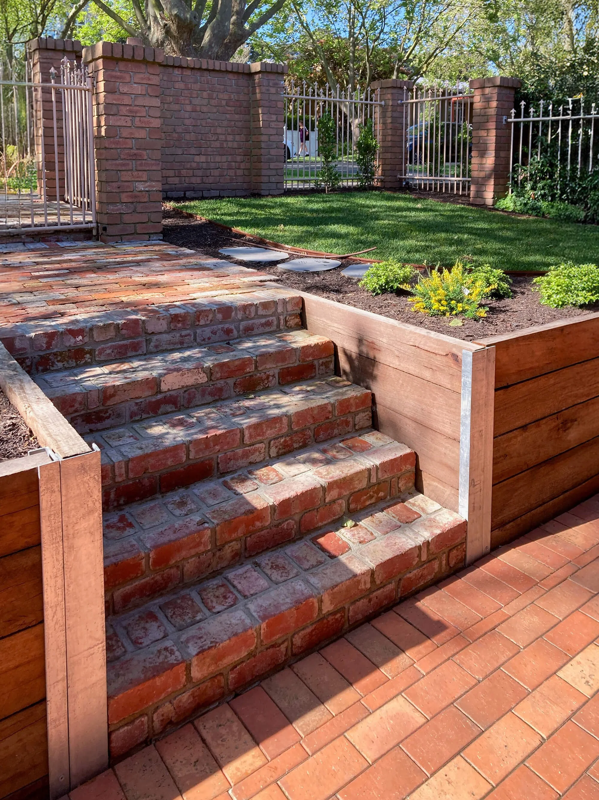 Brick steps leading up to a backyard garden with a brick wall and metal gate, surrounded by lush trees and plants.