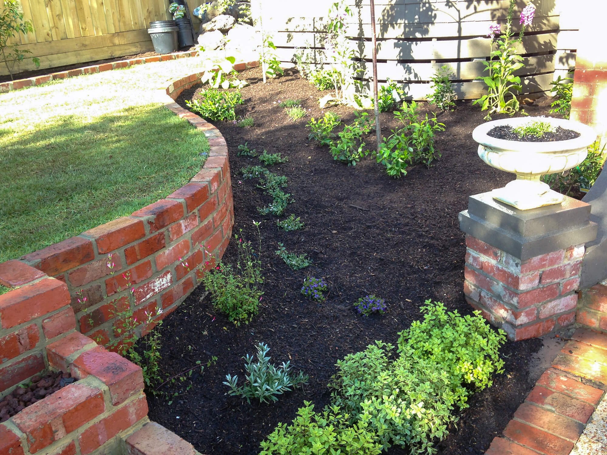 A garden with a brick curved border, dark soil, and a variety of green plants. There is a white decorative planter on a brick pillar in the right foreground, and a wooden fence with shadows in the background.