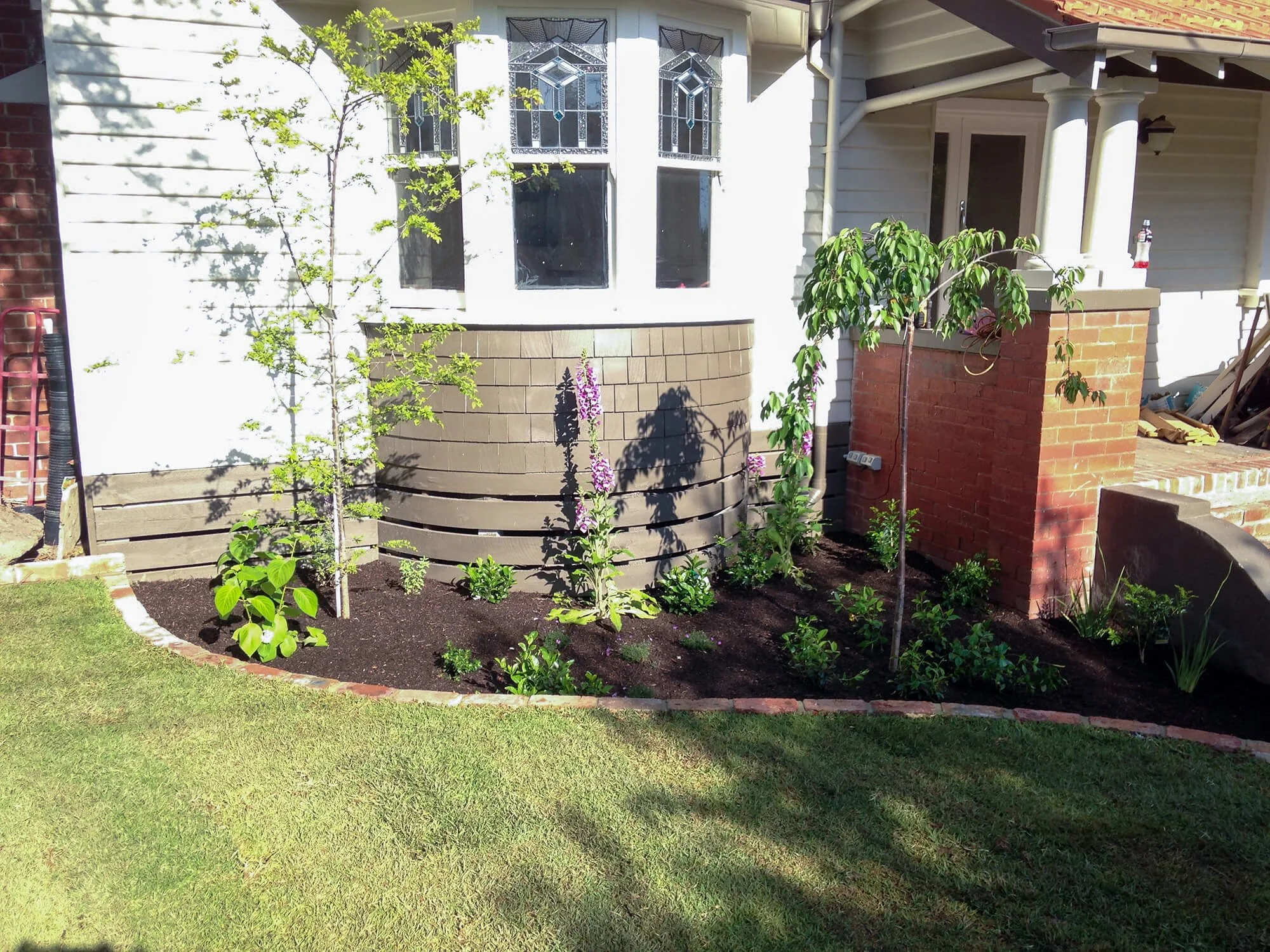 A small garden bed in front of a house with a white exterior, featuring young trees, flowering plants, and newly planted greenery, bordered by a brick edging.