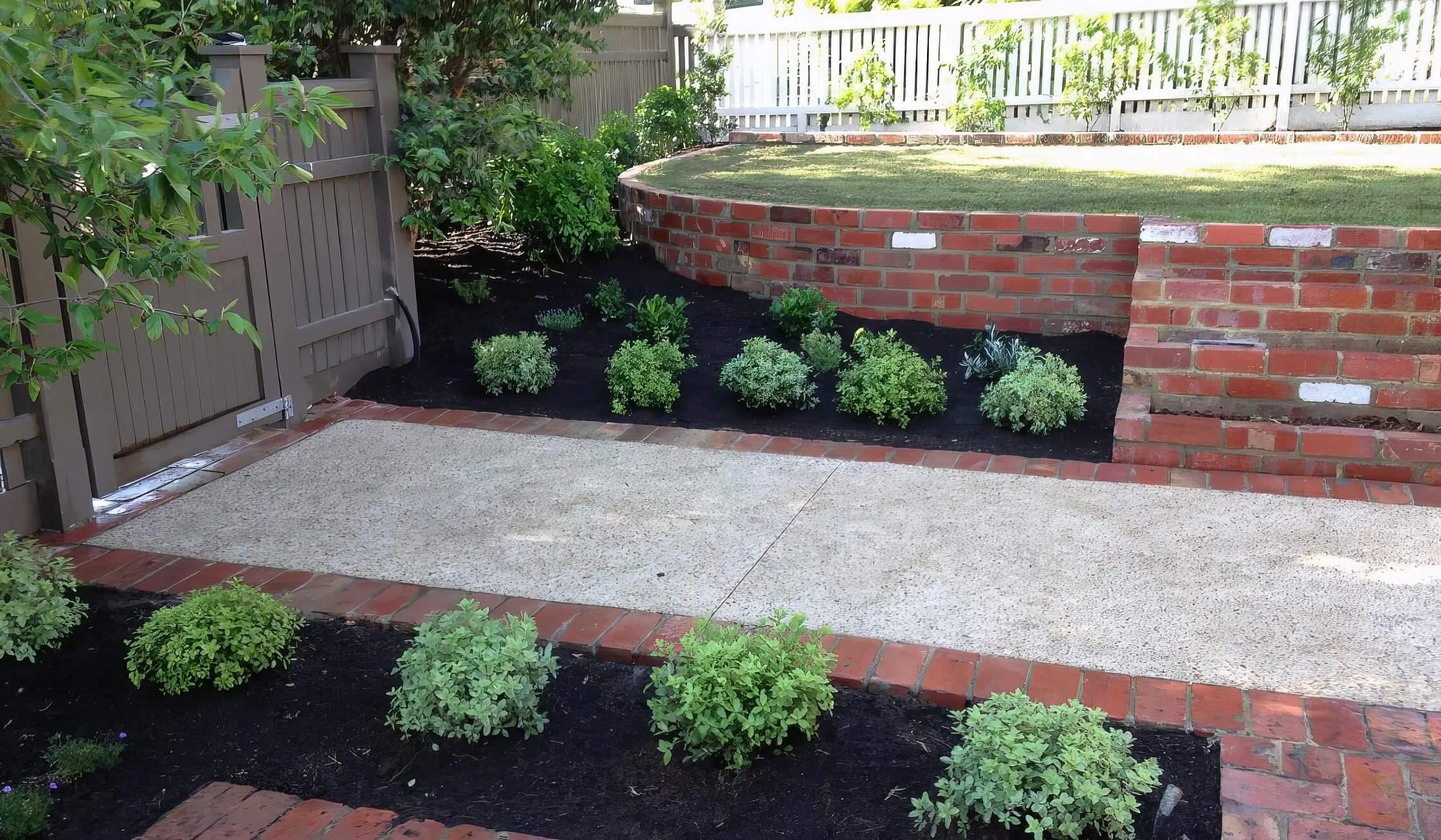 A backyard garden features a raised brick flower bed on the right side, with green plants and bushes. The area in the foreground has a concrete patio with a small section of brick border. A wooden gate and a fence surround the yard, and there are tre