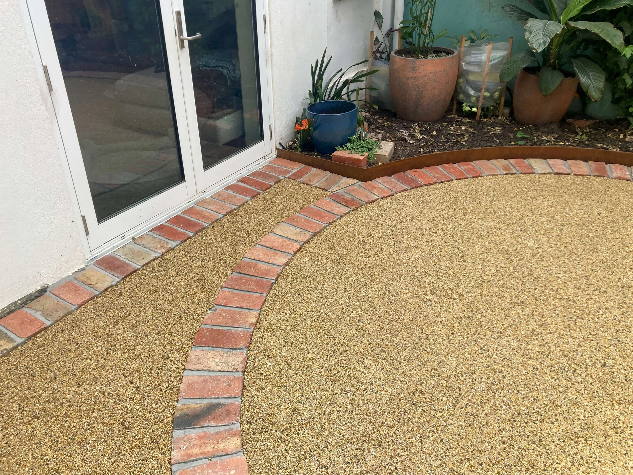 An outdoor patio area with a glass door, brick and pebble patterned walkway, and various potted plants and greenery.