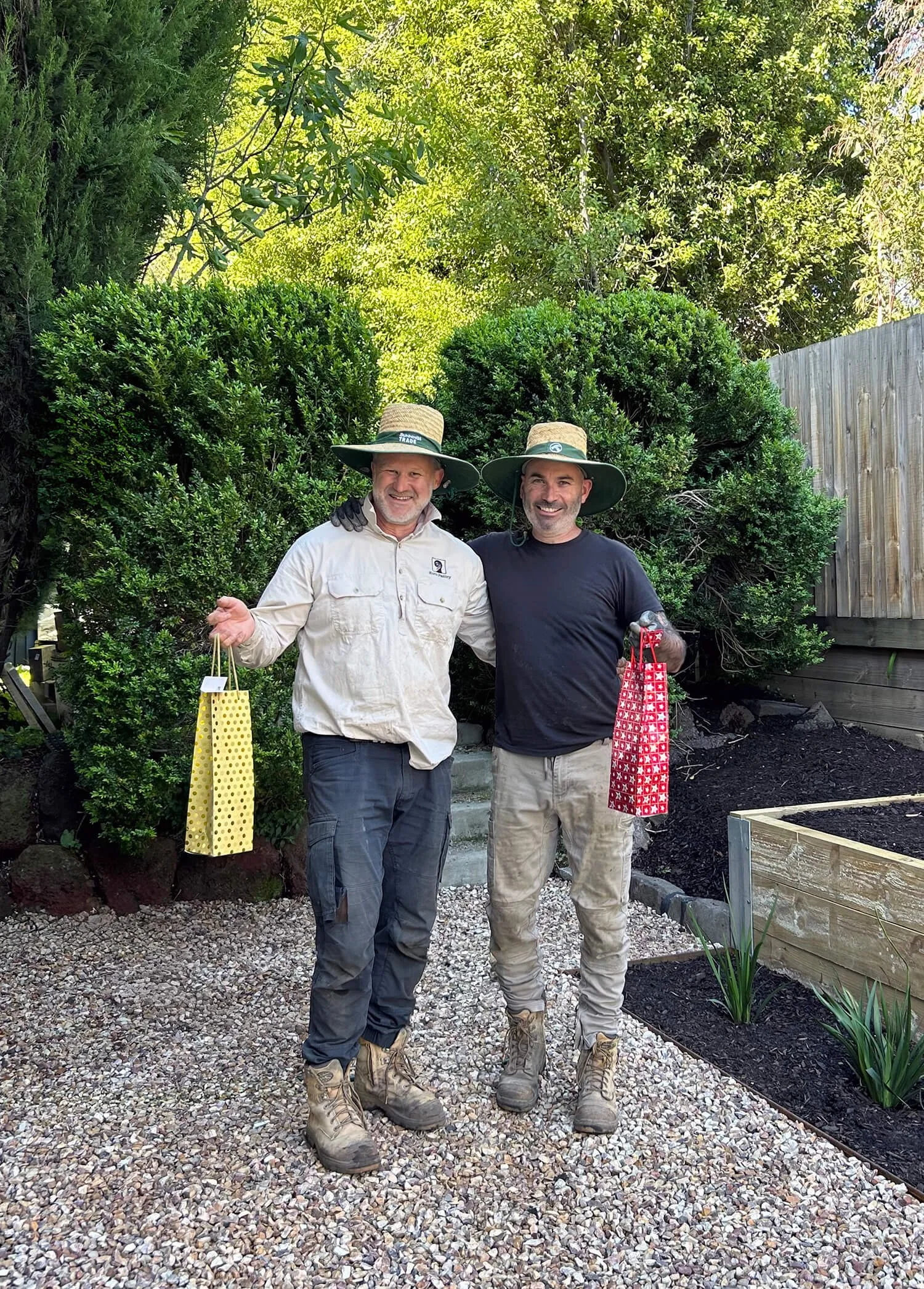 Two men stand outdoors in a garden, smiling, wearing wide-brimmed hats, casual clothes, and boots. They are holding gift bags, with greenery and trees in the background.