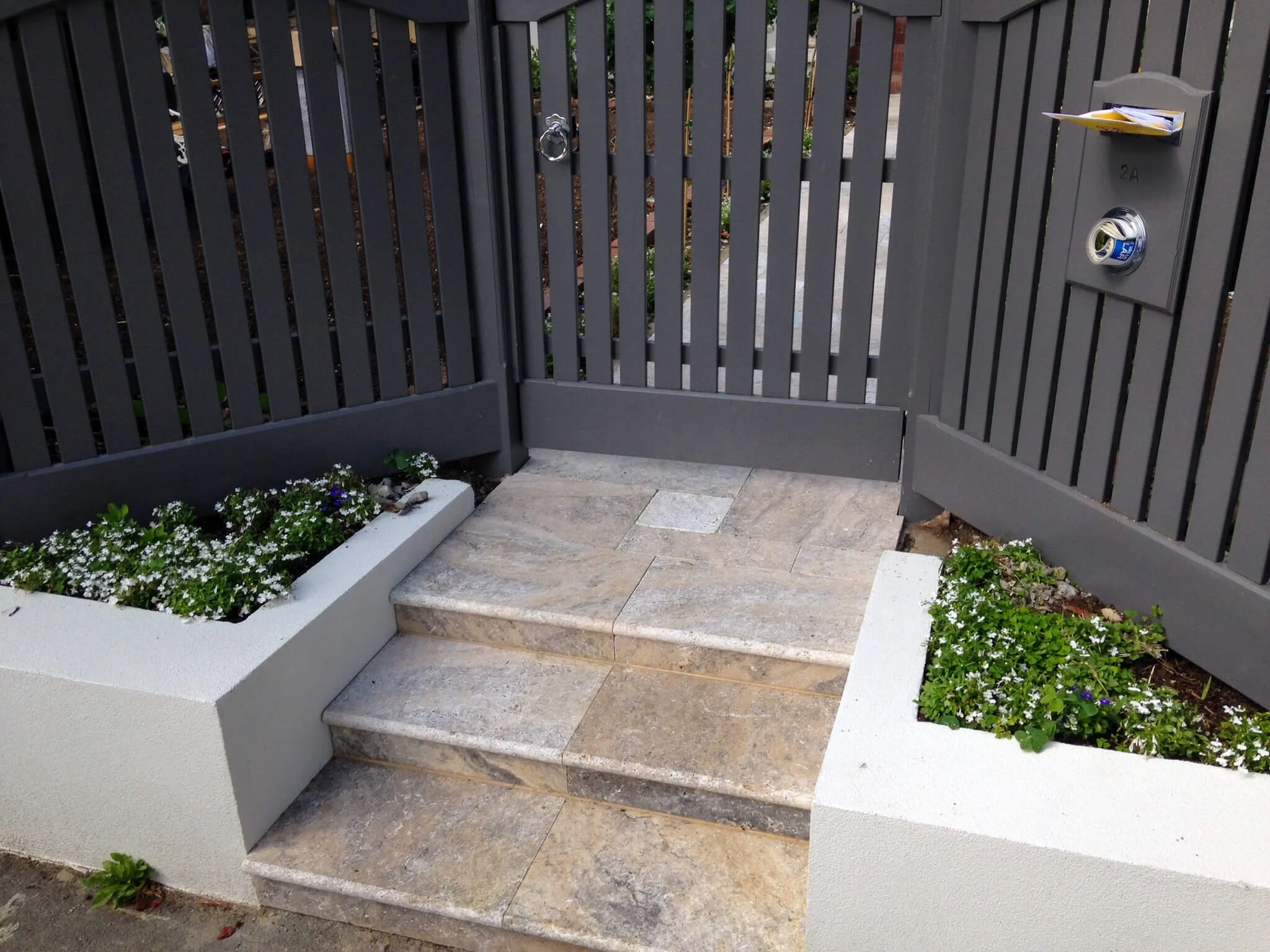 A modern gray metal gate with vertical slats, a lock, and a mailbox, flanked by concrete planters with white and purple flowers, leading to a front yard with a pathway and greenery.